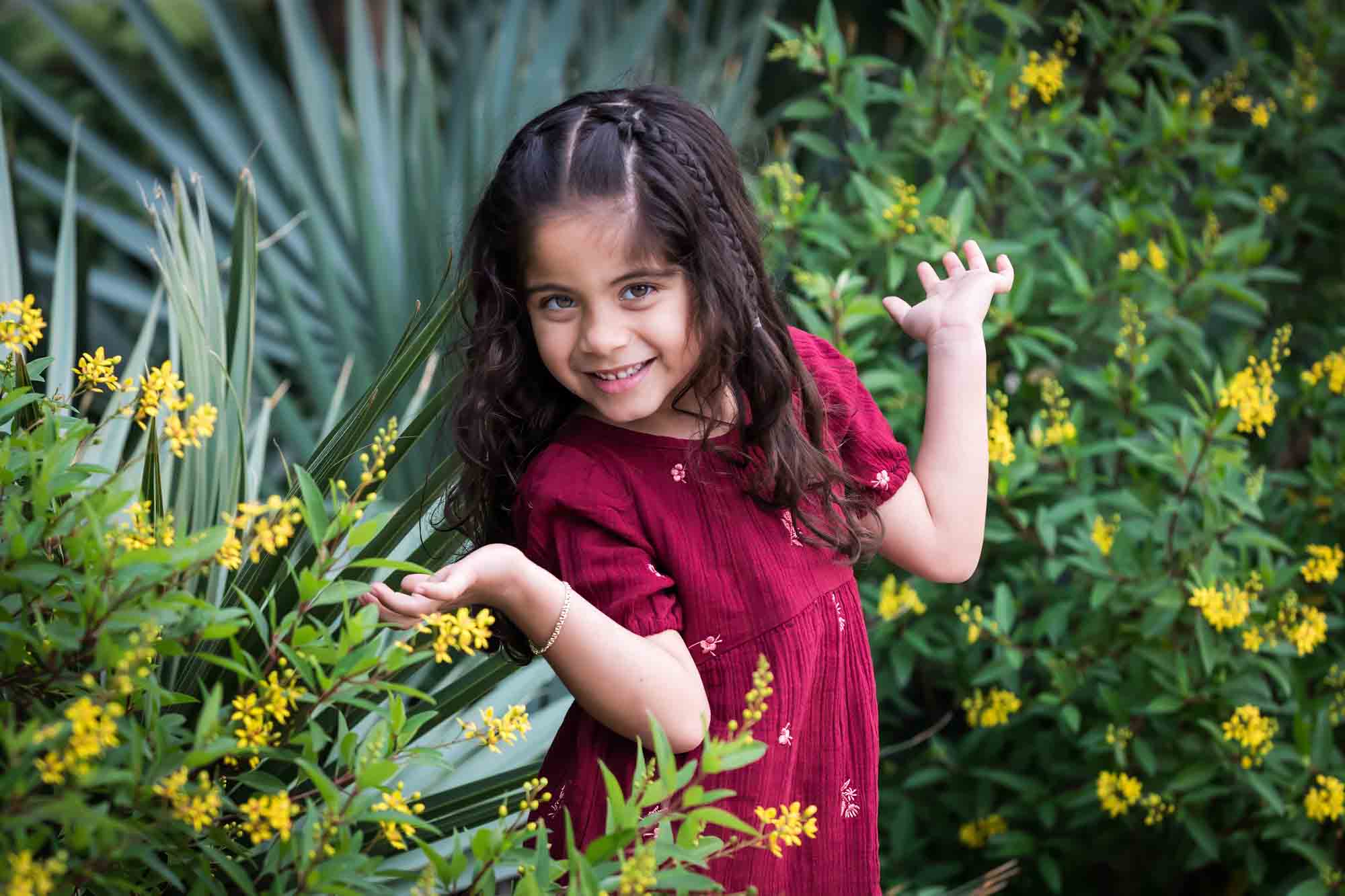 Little girl with long brown hair wearing maroon dress standing in front of green bushes