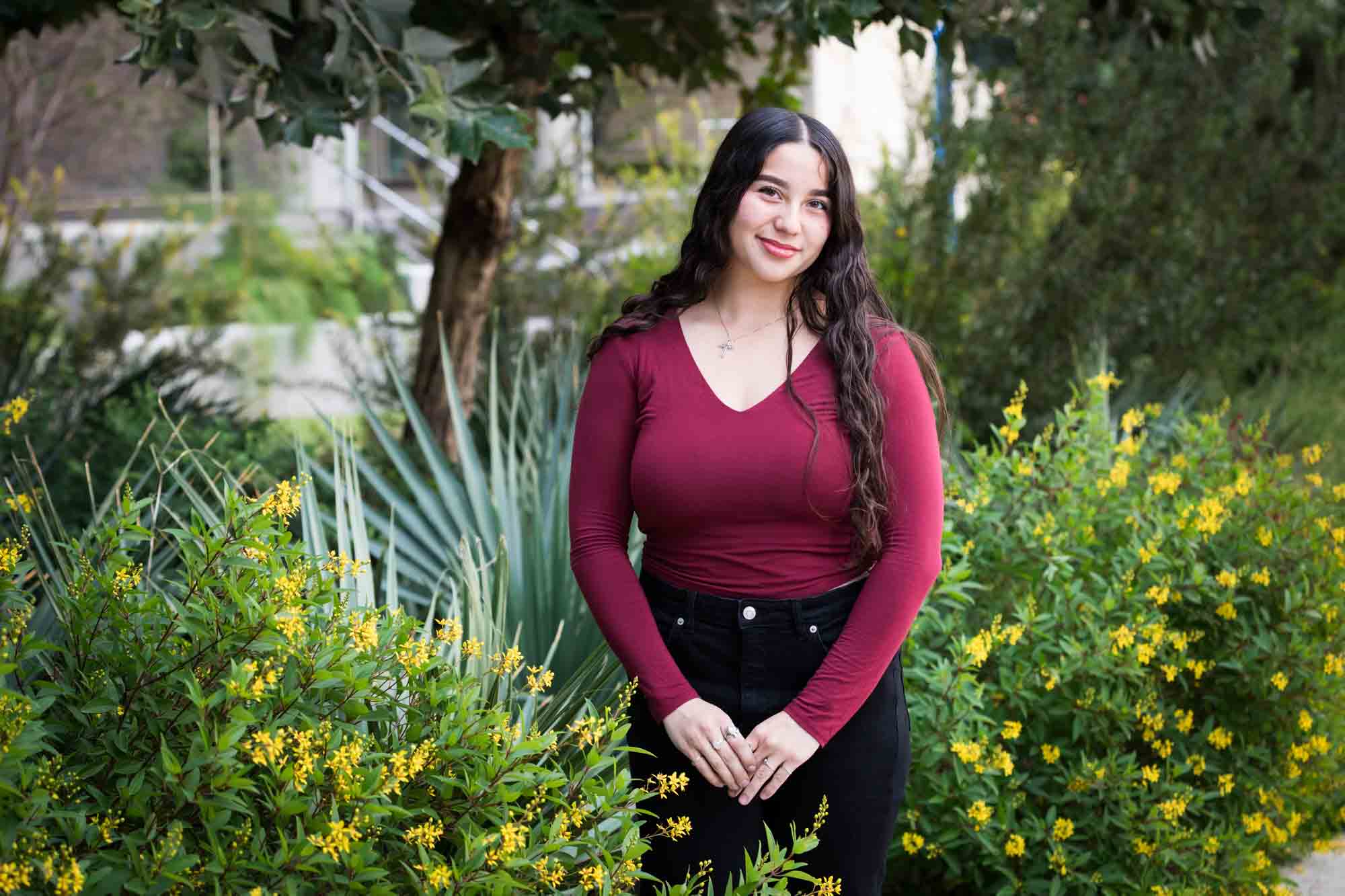 Teen girl with long brown hair wearing maroon top standing in front of green bushes