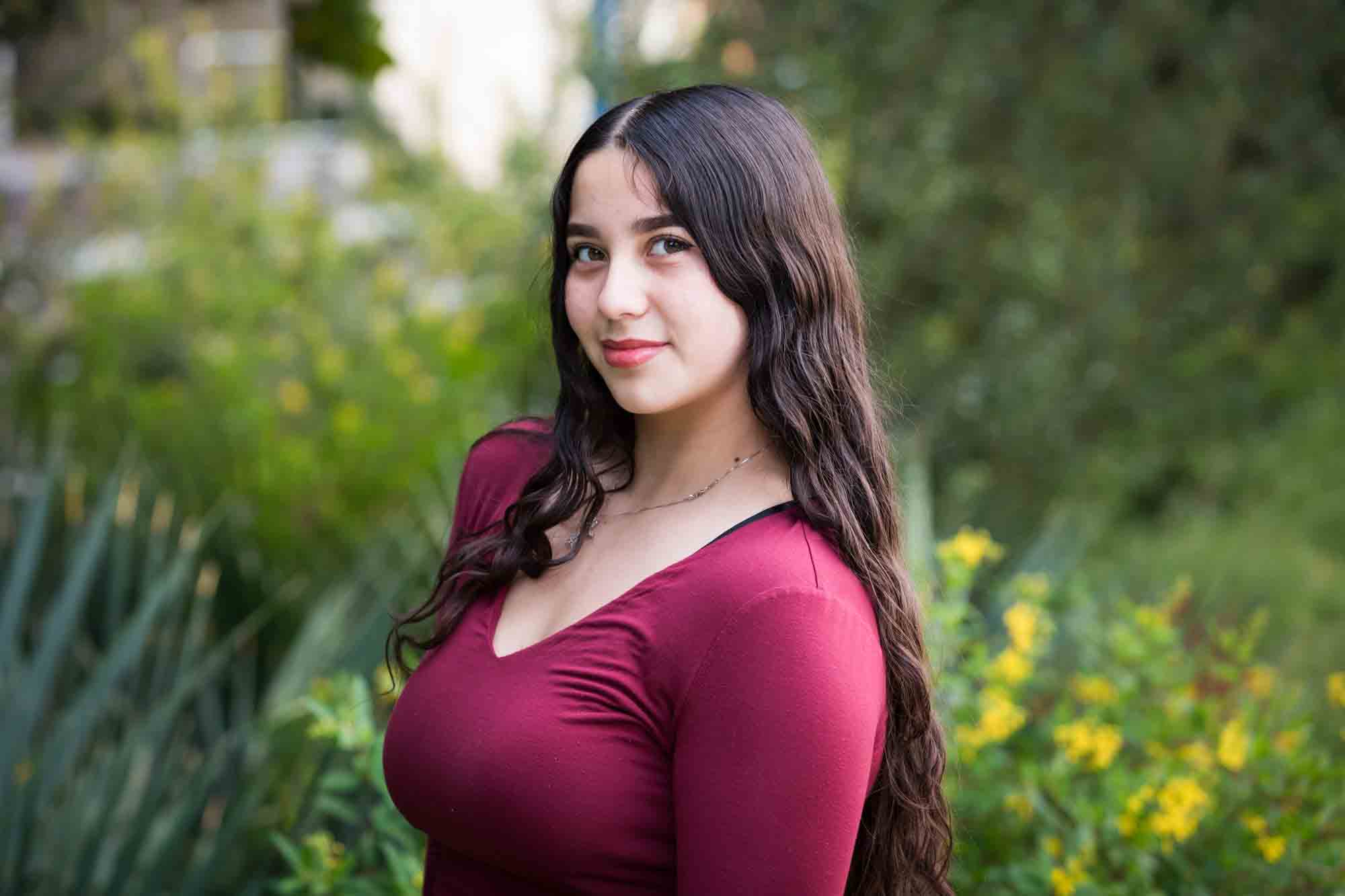 Teen girl with long brown hair wearing maroon top standing in front of green bushes