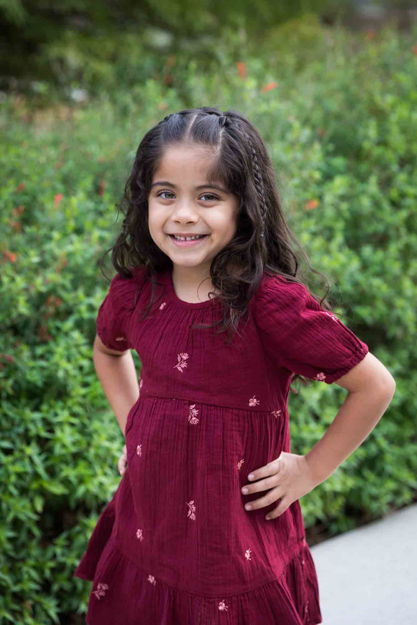 Little girl with long brown hair wearing maroon dress standing in front of green bushes with hands on hips during a San Pedro Creek Park family portrait session