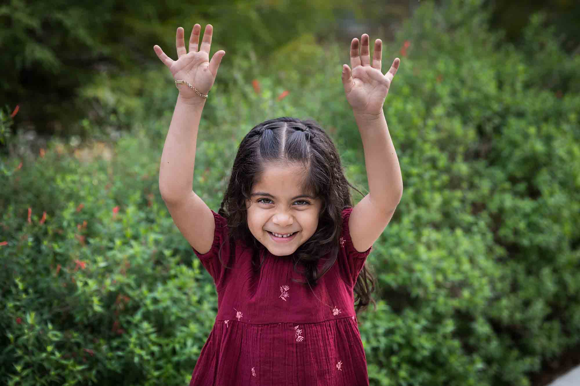 Little girl with long brown hair wearing maroon dress standing in front of green bushes with hands outstretched during a San Pedro Creek Park family portrait session