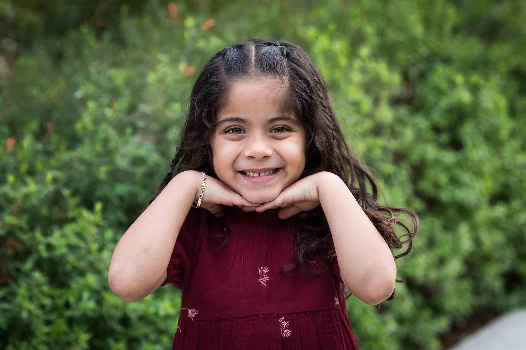 Little girl with long brown hair wearing maroon dress standing in front of green bushes with hands under chin during a San Pedro Creek Park family portrait session