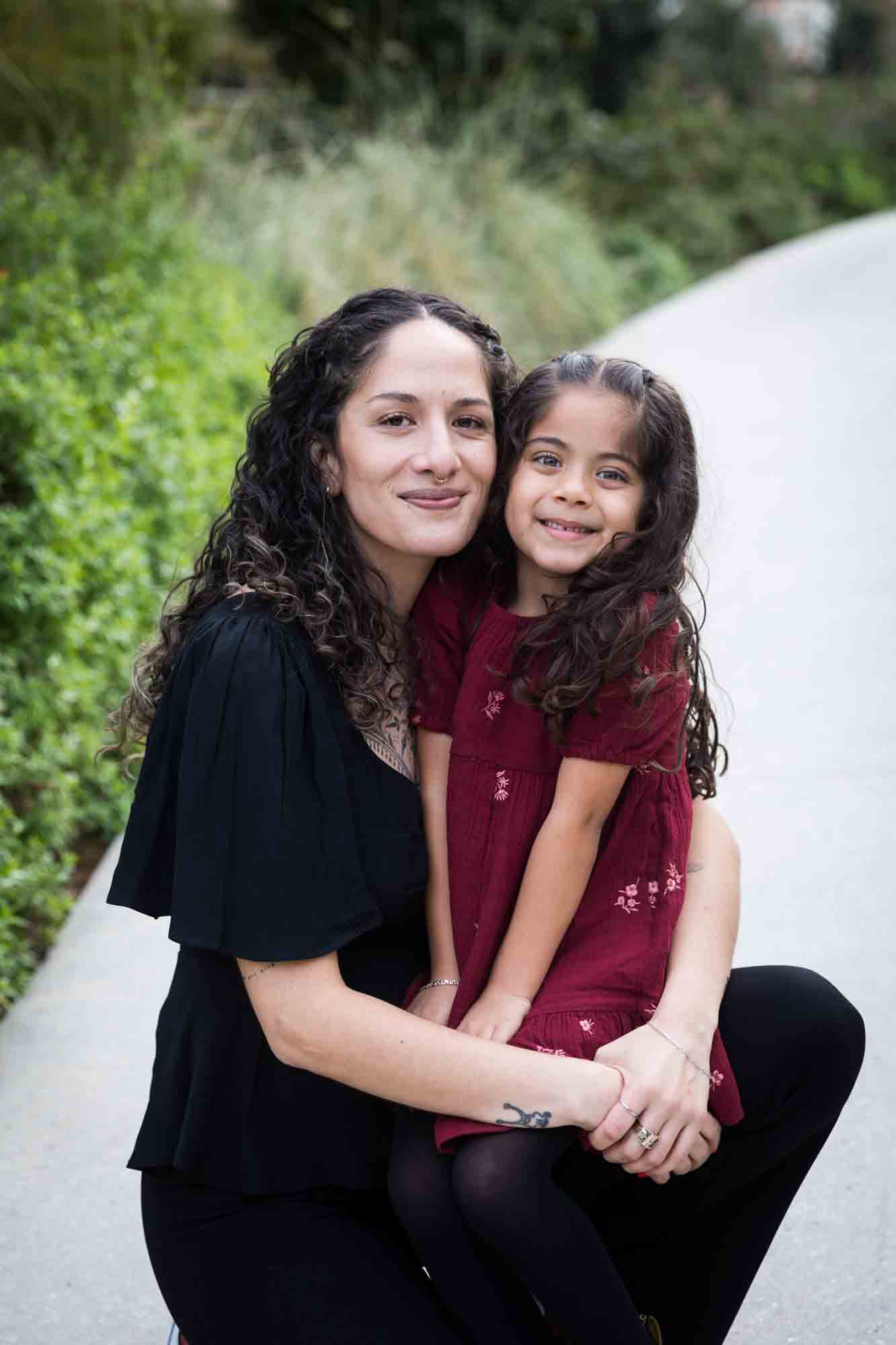 Mother and daughter hugging on sidewalk in front of green bushes during a San Pedro Creek Park family portrait session