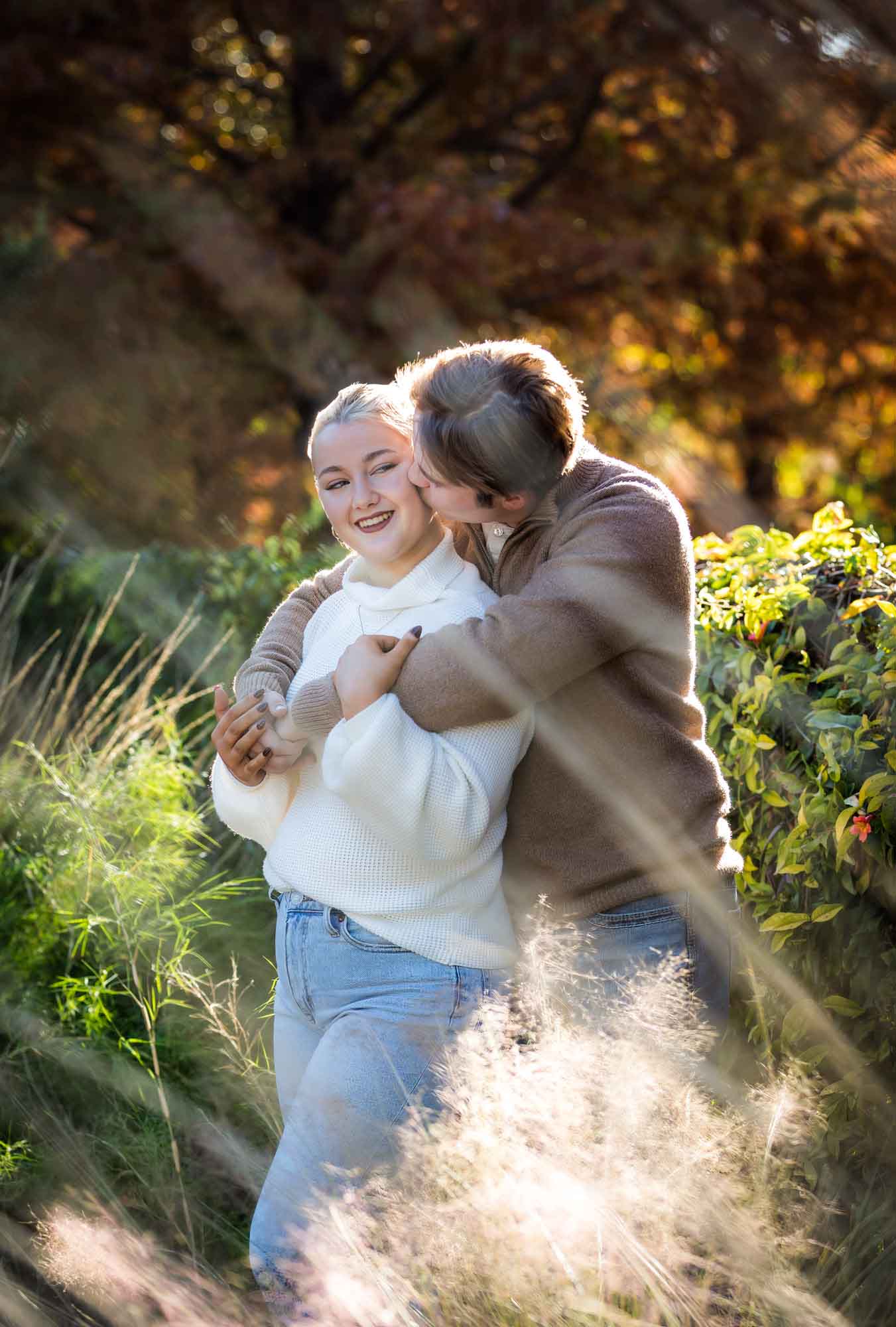 Couple wearing sweaters and hugging in front of colorful bushes at the Pearl during an engagement photo shoot