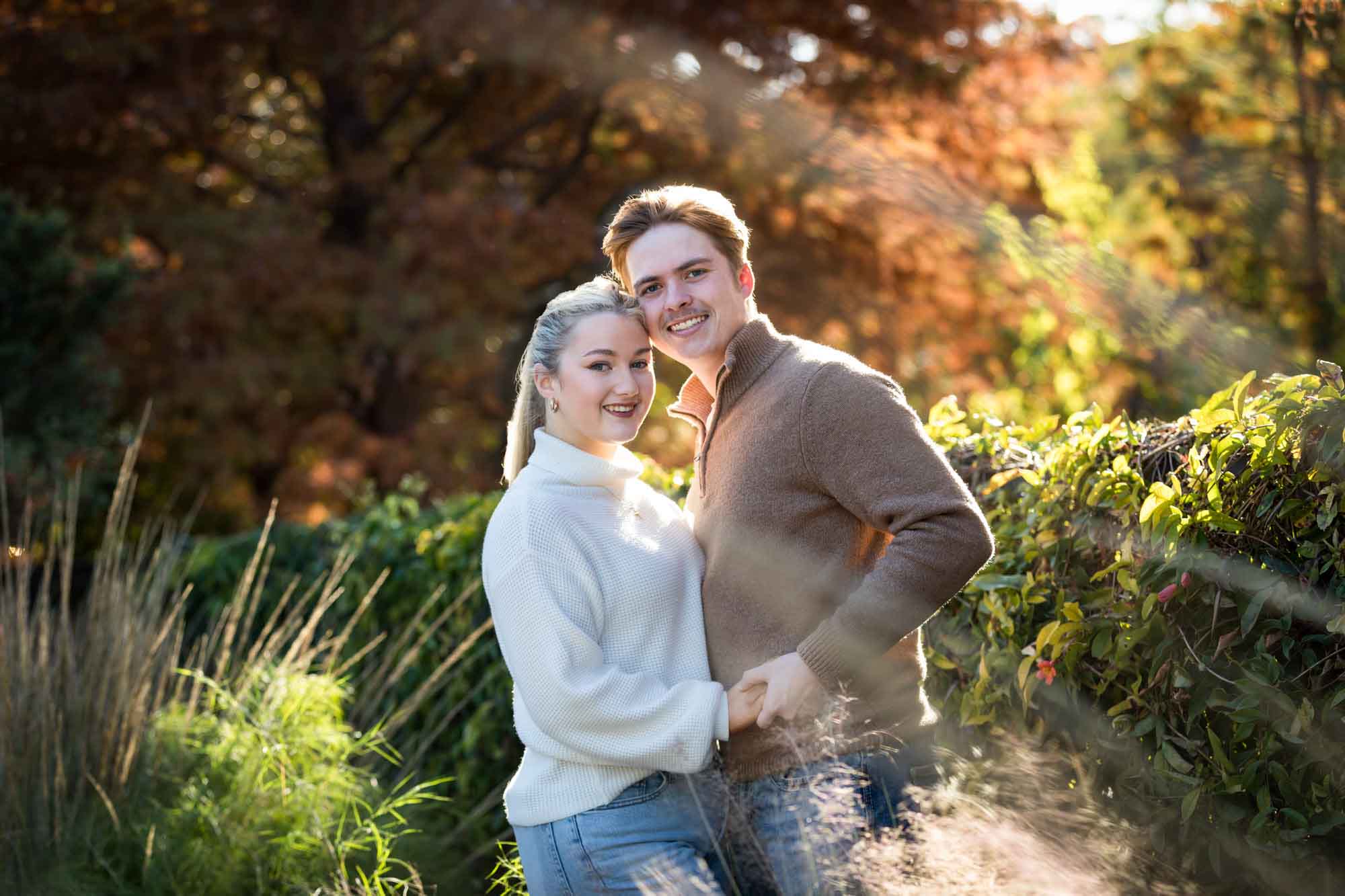 Couple wearing sweaters and hugging in front of colorful bushes at the Pearl during an engagement photo shoot