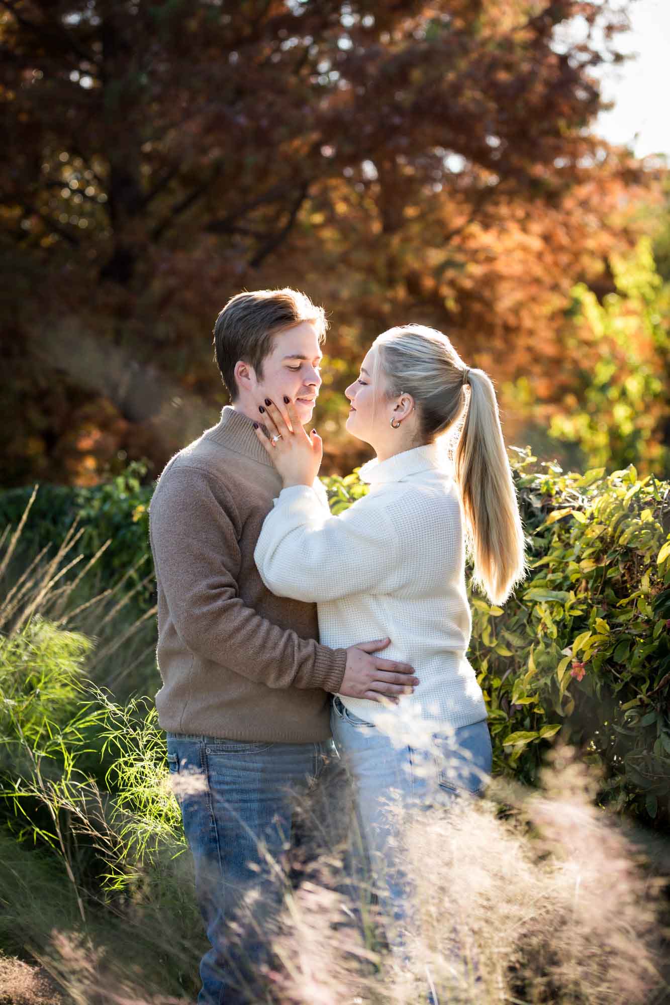 Couple wearing sweaters and hugging in front of colorful bushes at the Pearl during an engagement photo shoot