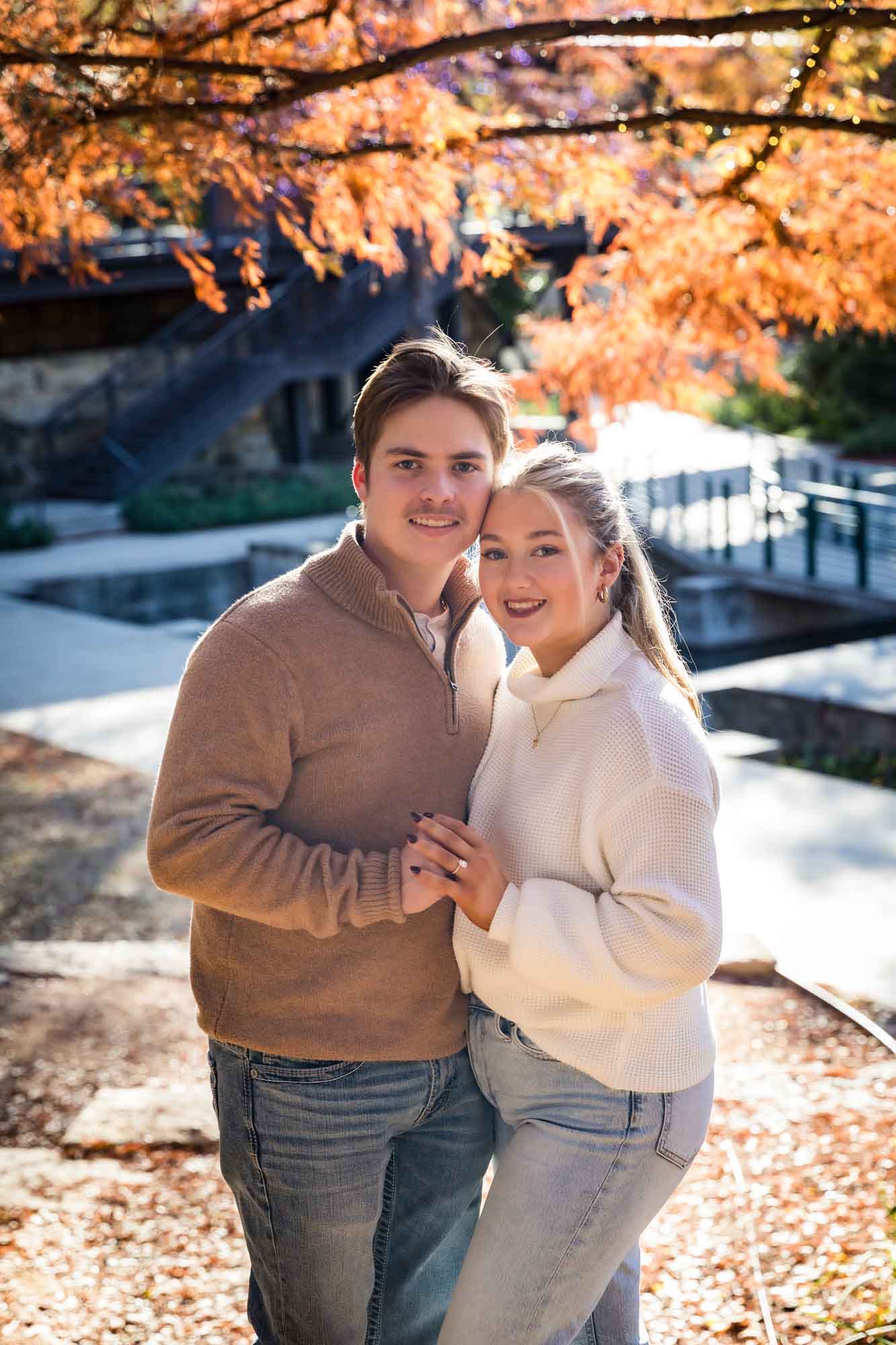 Couple wearing sweaters and hugging in front of colorful bushes at the Pearl during an engagement photo shoot