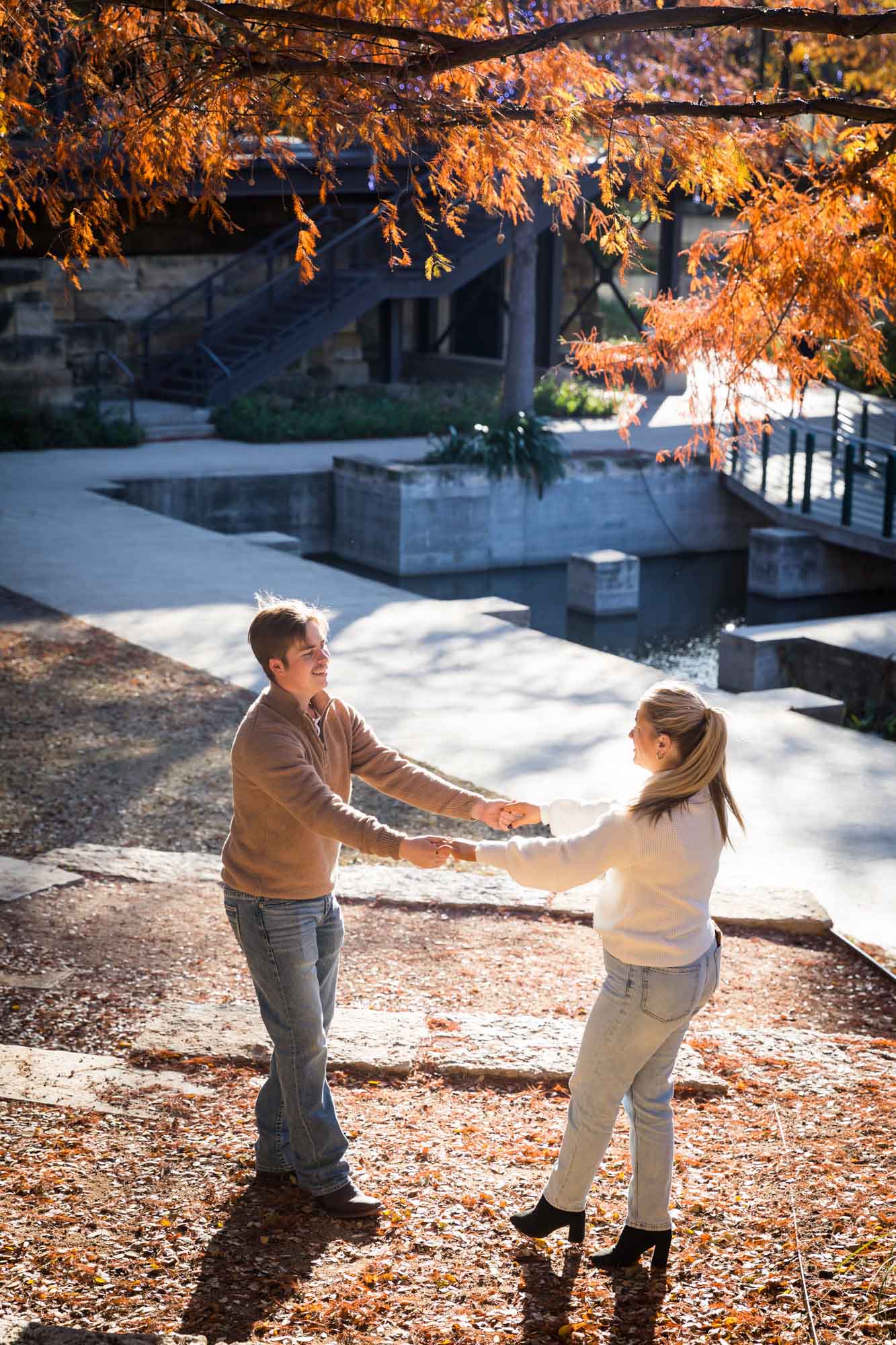 Couple wearing sweaters and dancing in front of the Riverwalk at the Pearl during an engagement photo shoot