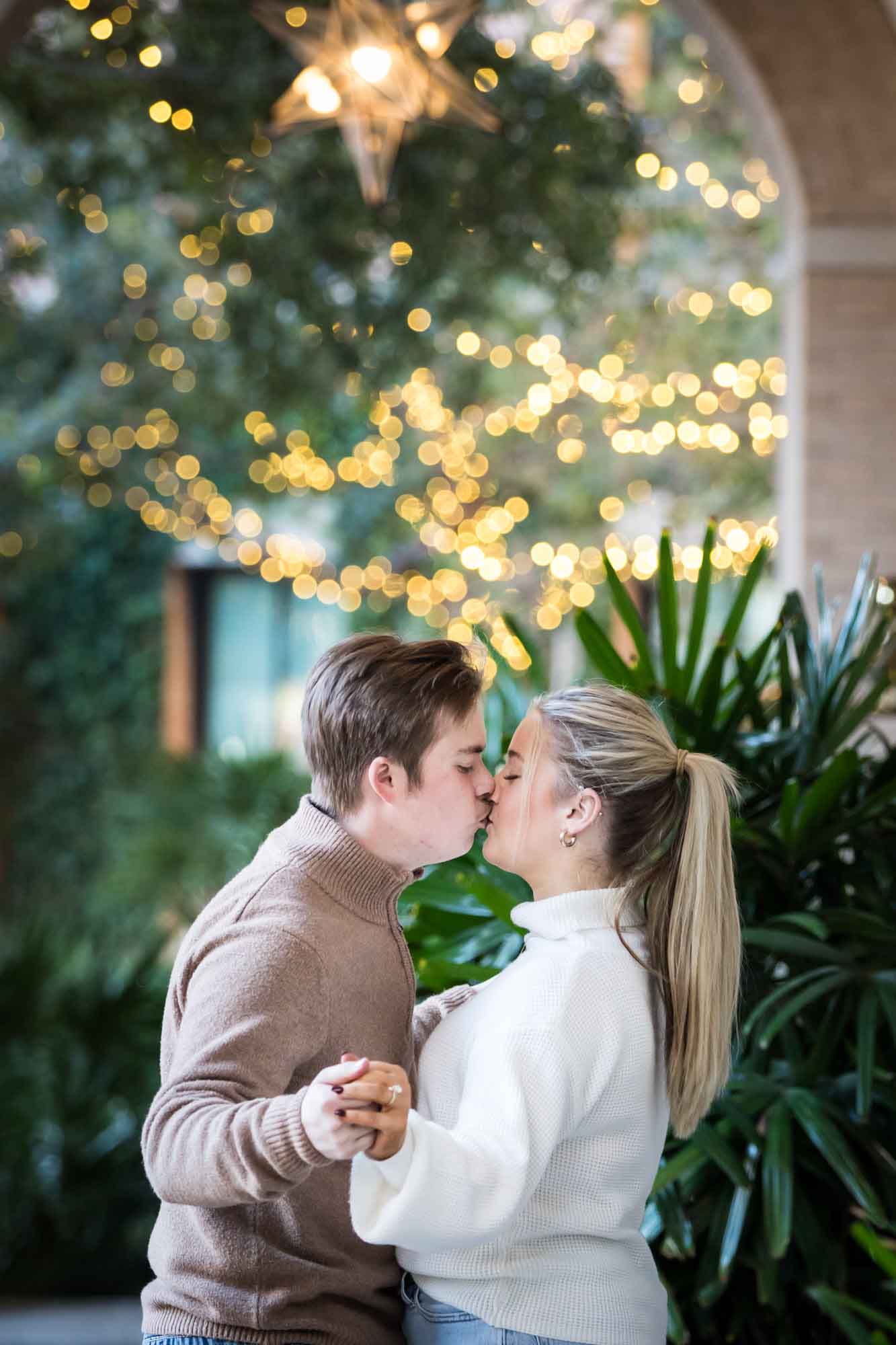 Couple wearing sweaters and kissing in front of string lights at the Pearl during an engagement photo shoot