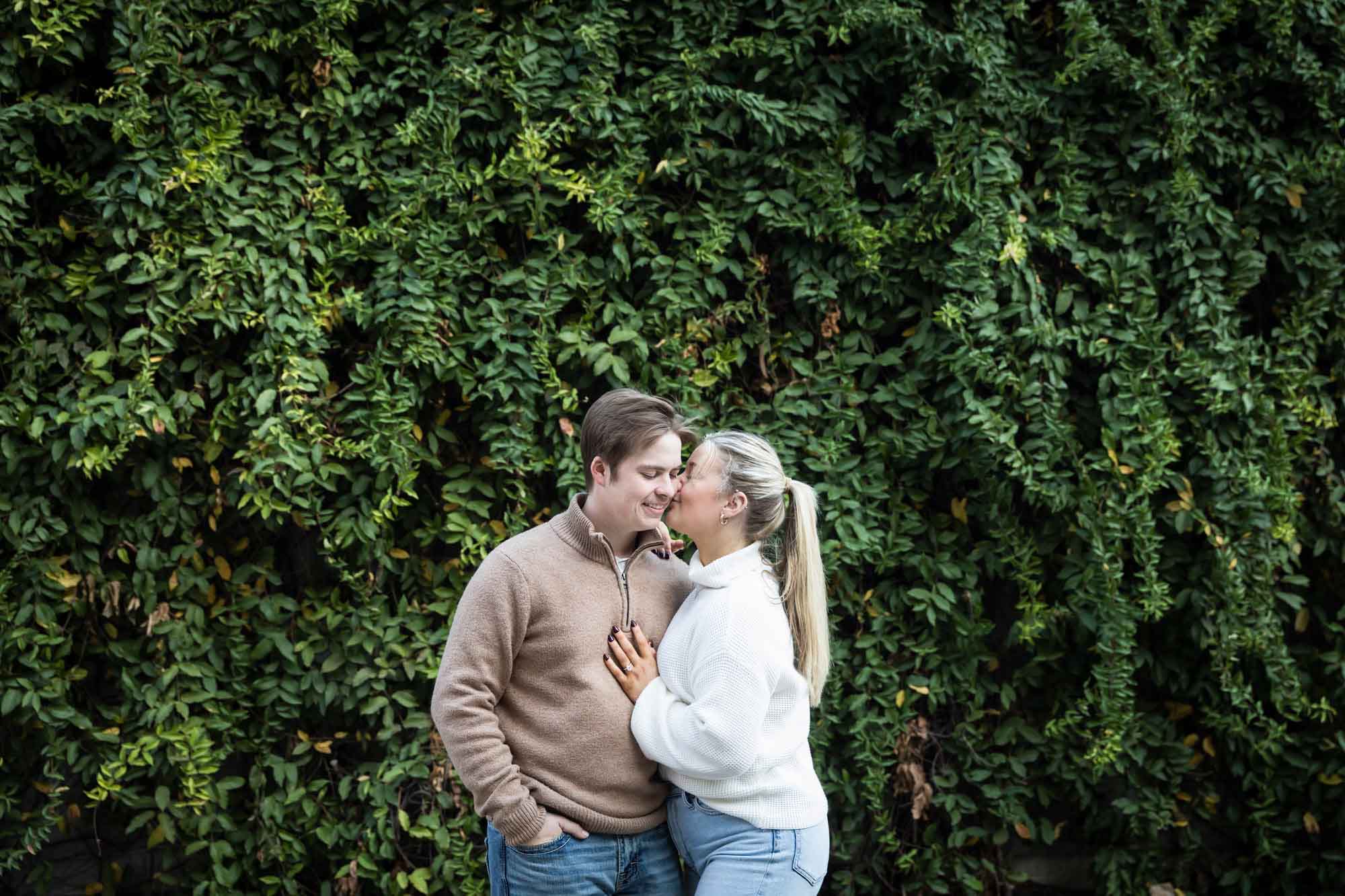 Couple wearing sweaters and kissing in front of wall of green ivy