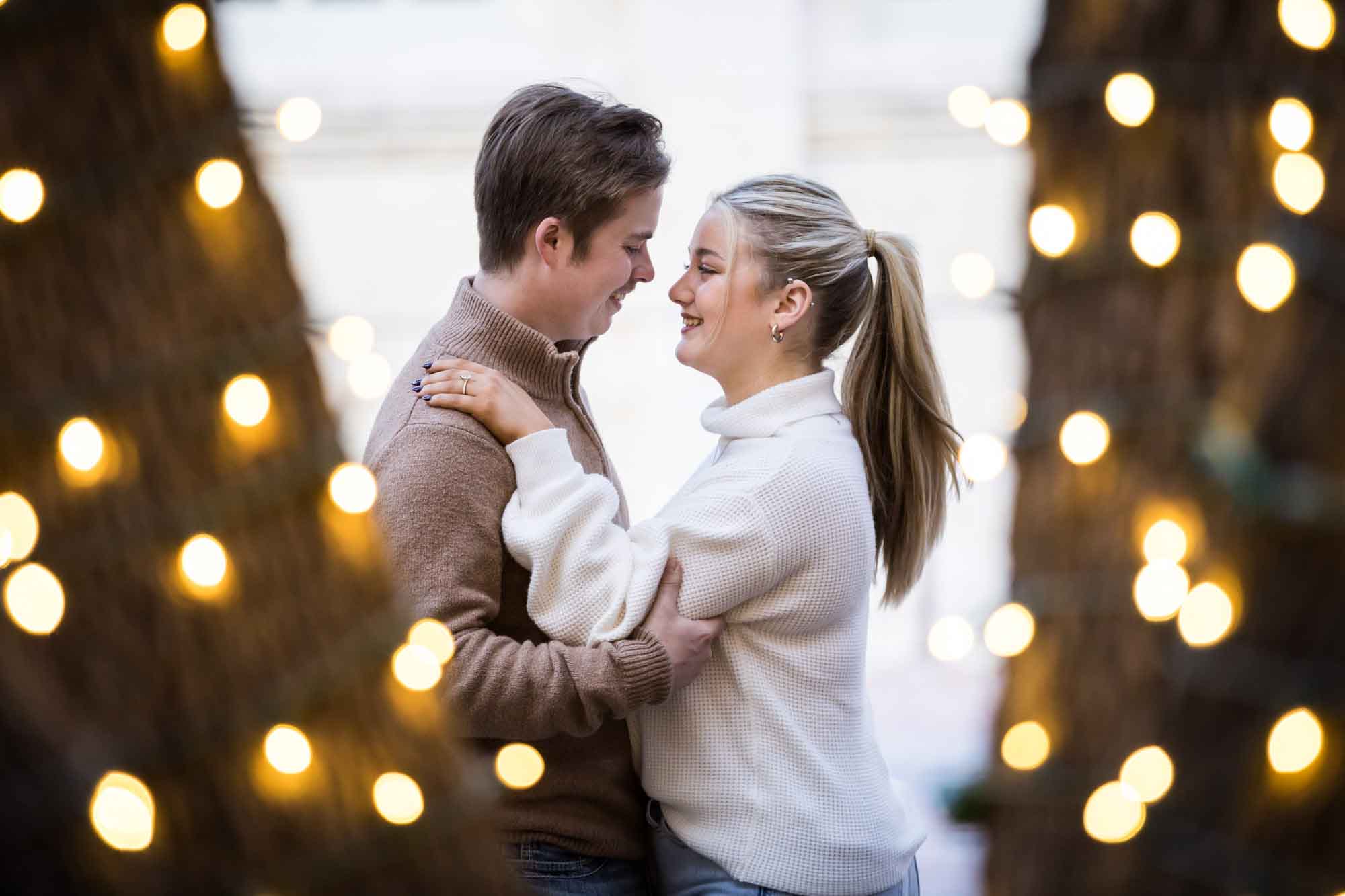 Couple hugging as viewed through tree wrapped in string lights for an article on the importance of timing for a surprise proposal