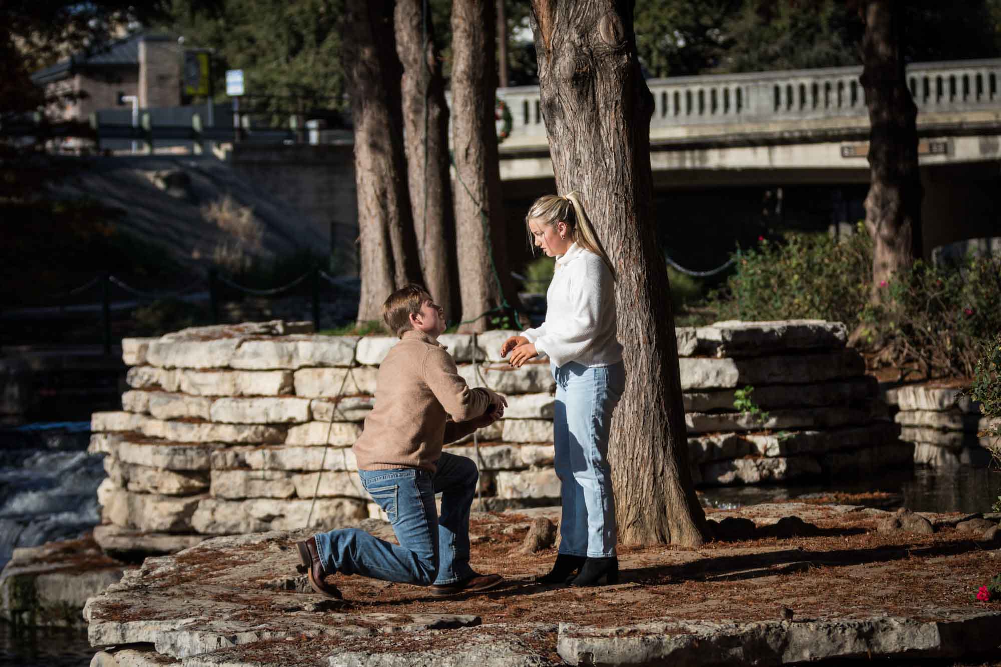 Man on one knee proposing to woman wearing white sweter for an article on the importance of timing for a surprise proposal