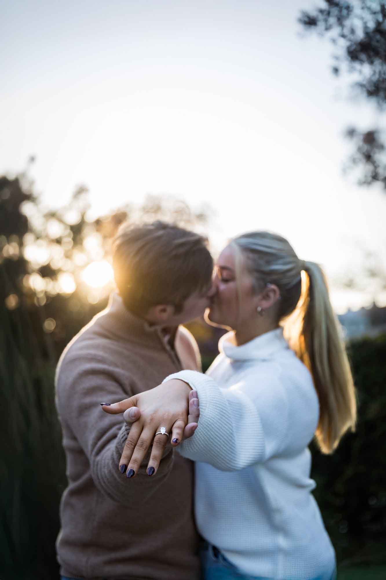 Couple kissing at sunset while throwing hand with engagement ring to camera