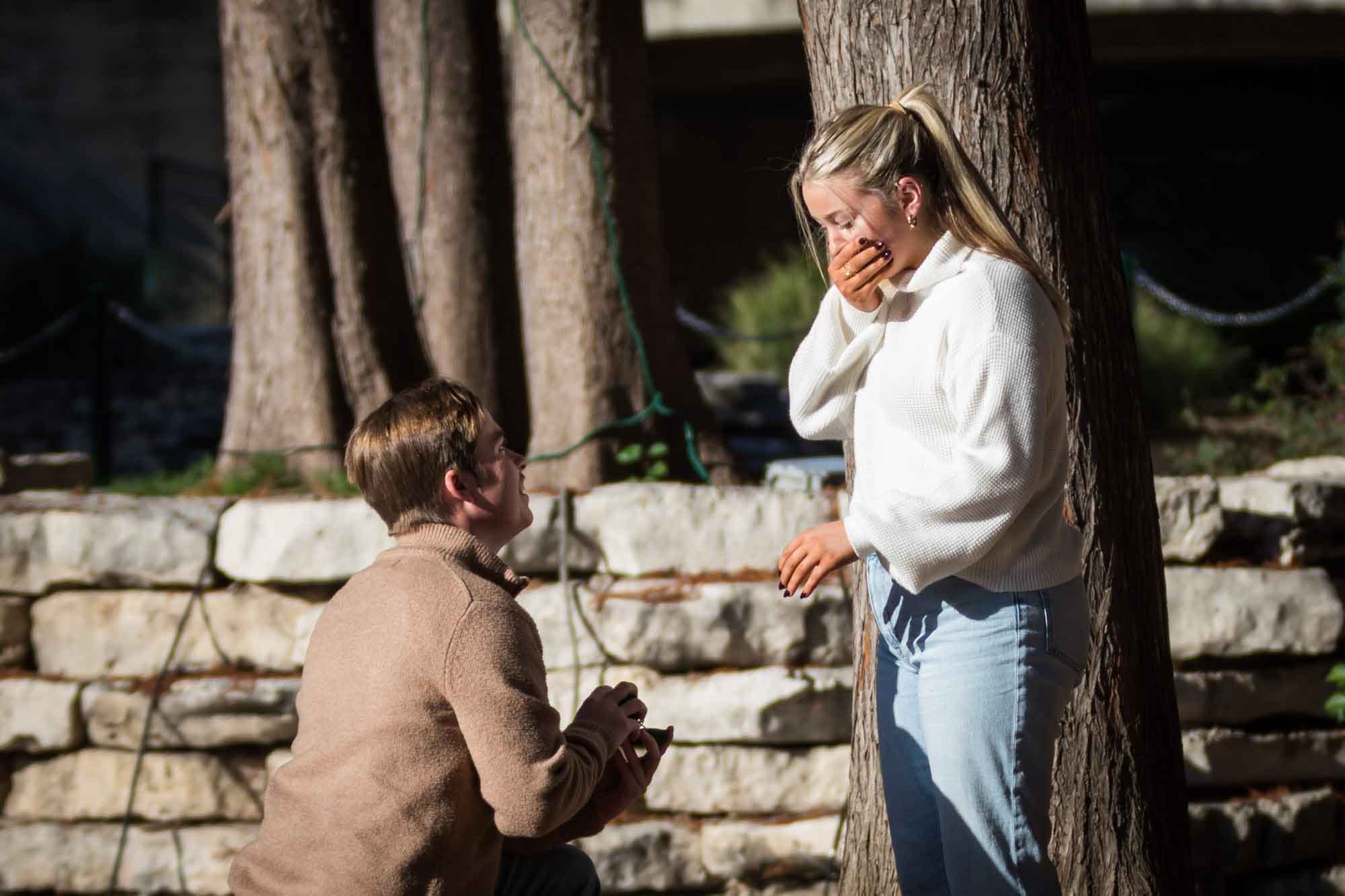 Man bending down proposing to woman wearing white sweater for an article on the importance of timing for a surprise proposal