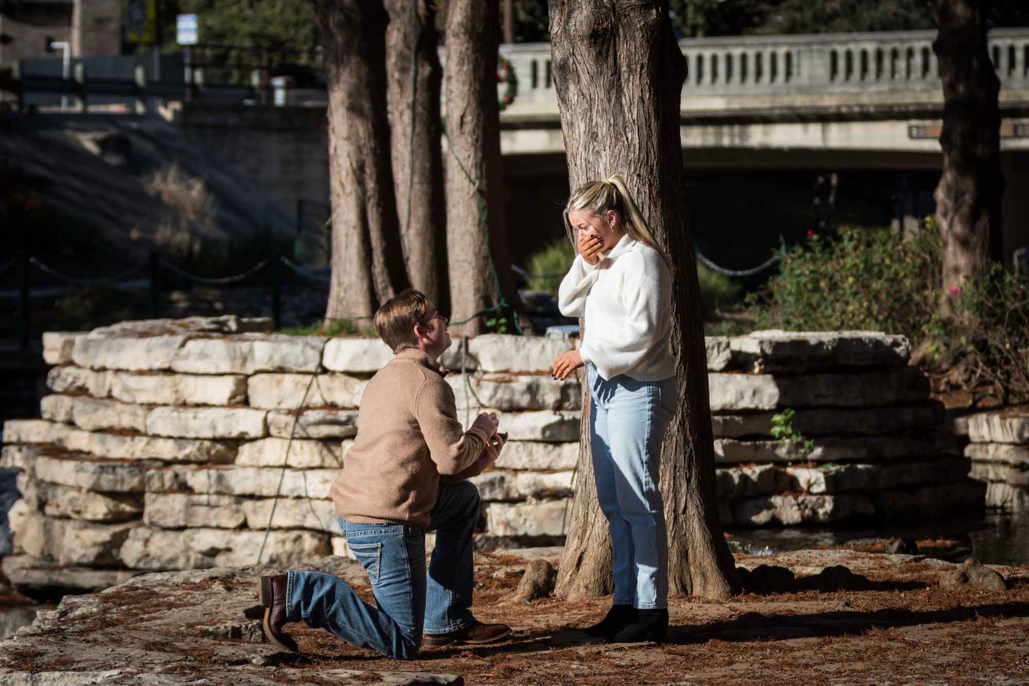 Man bending down proposing down to woman wearing white sweater for an article on the importance of timing for a surprise proposal