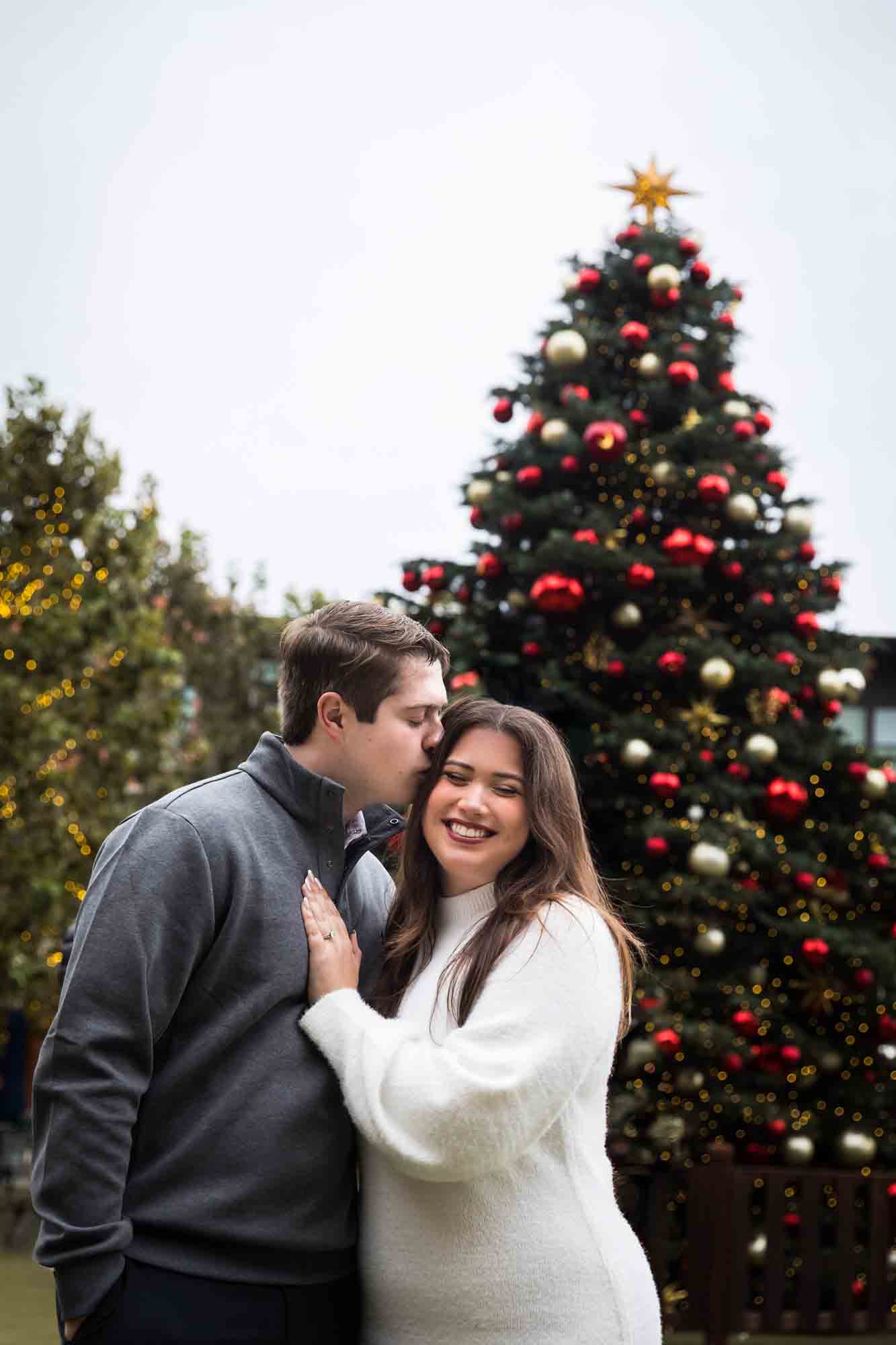 Man kissing woman on cheek in front of Christmas tree during an engagement photo shoot at the Pearl