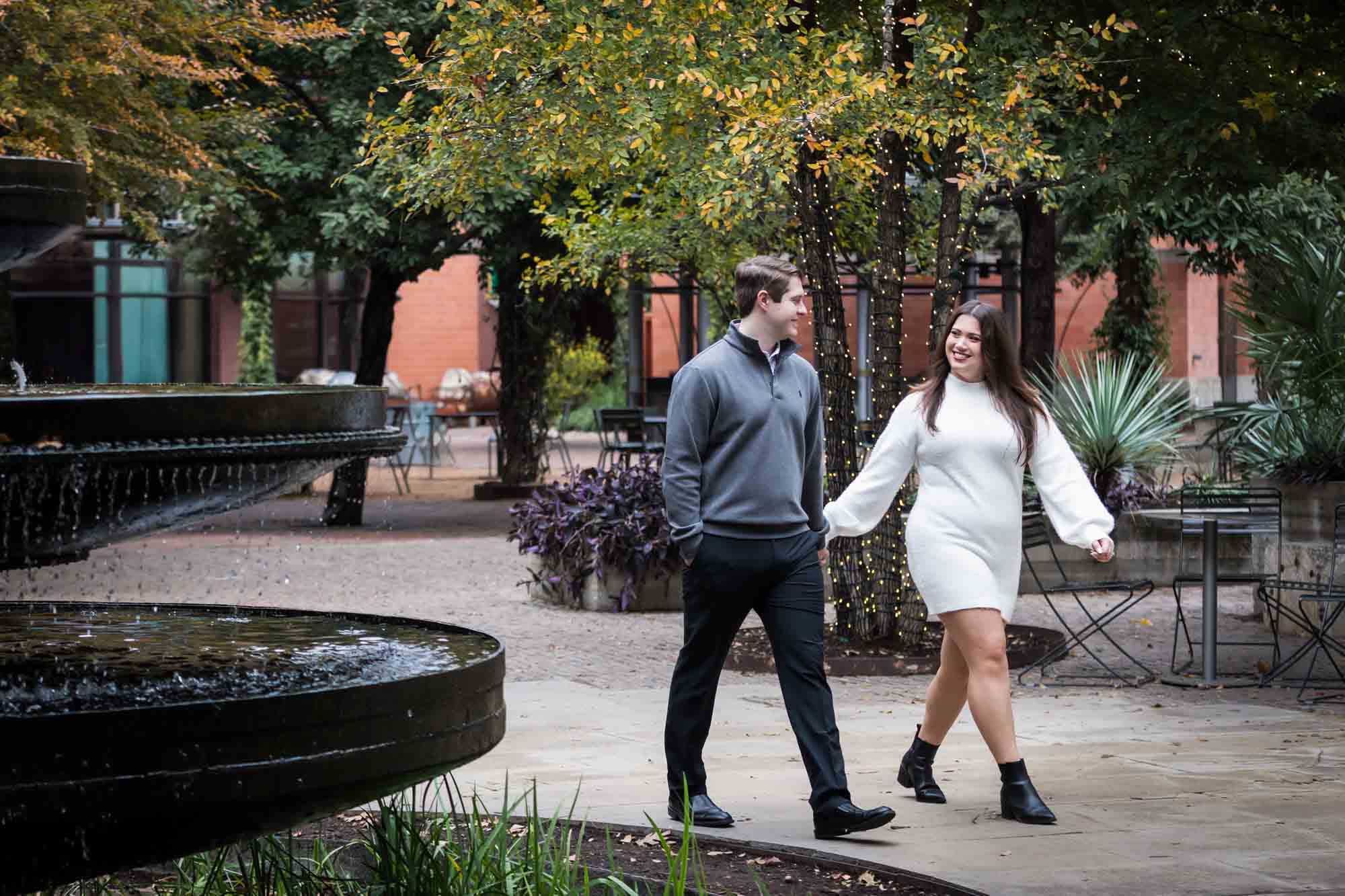 Couple walking past fountain and trees wrapped in string lights during a surprise proposal at the Pearl