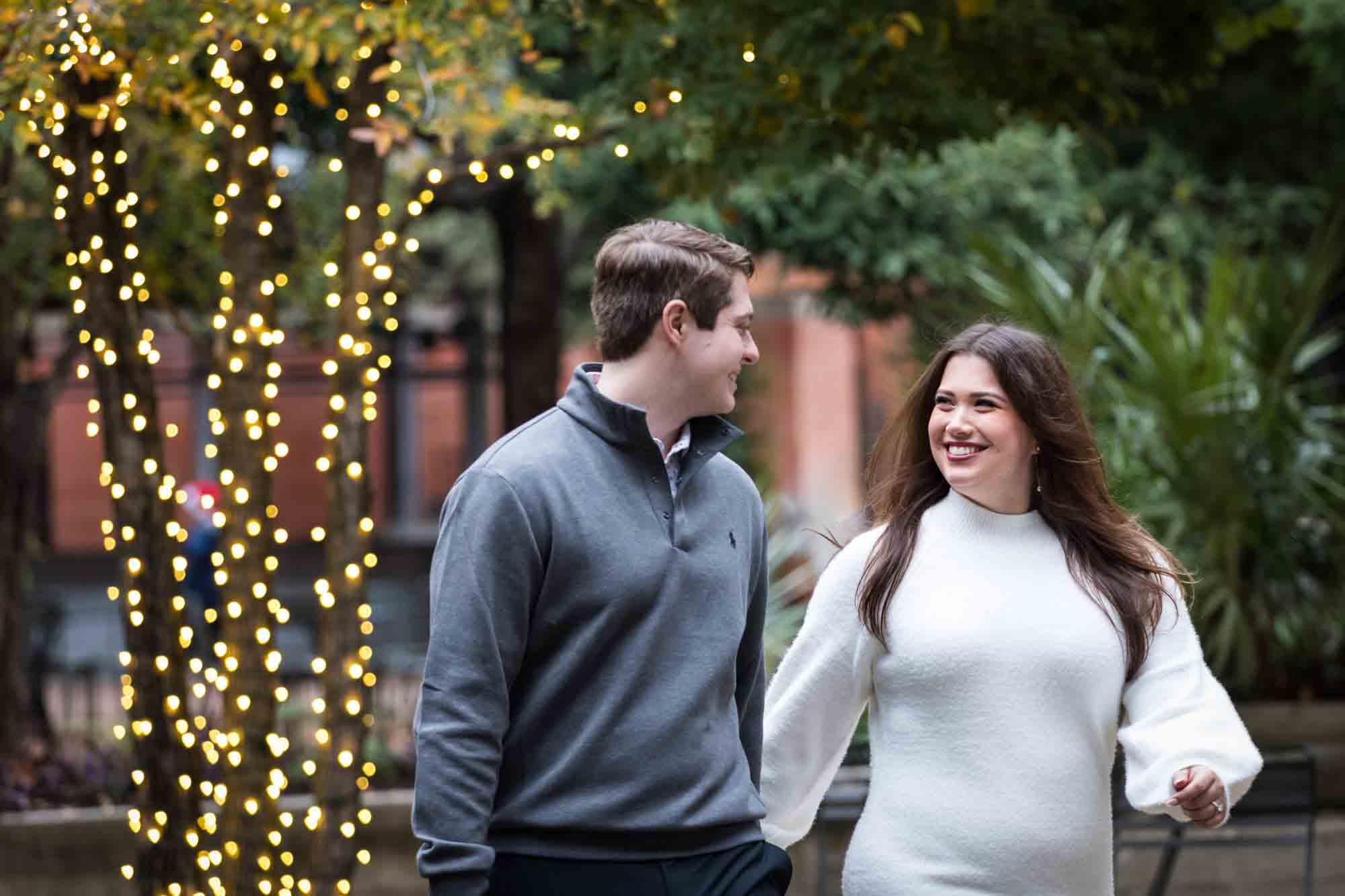 Couple walking in front of trees wrapped in string lights during a surprise proposal at the Pearl