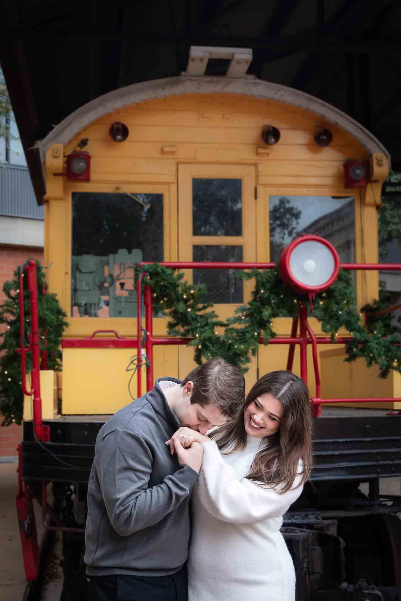 Man kissing woman's hand in front of yellow train trolley during a surprise proposal at the Pearl