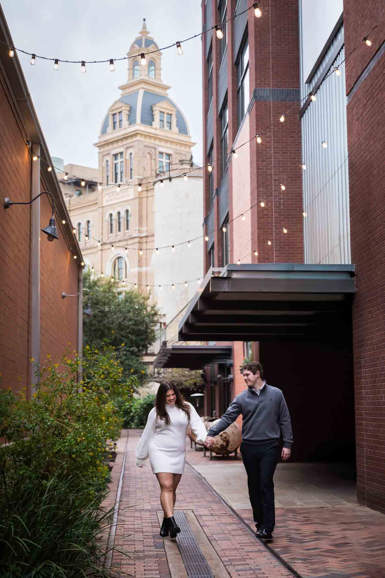 Couple walking down alleyway with Historic Pearl building in background during an engagement photo shoot at the Pearl