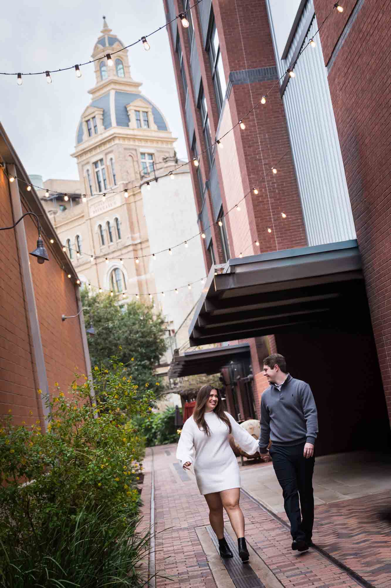 Couple walking down alleyway with Historic Pearl building in background during an engagement photo shoot at the Pearl