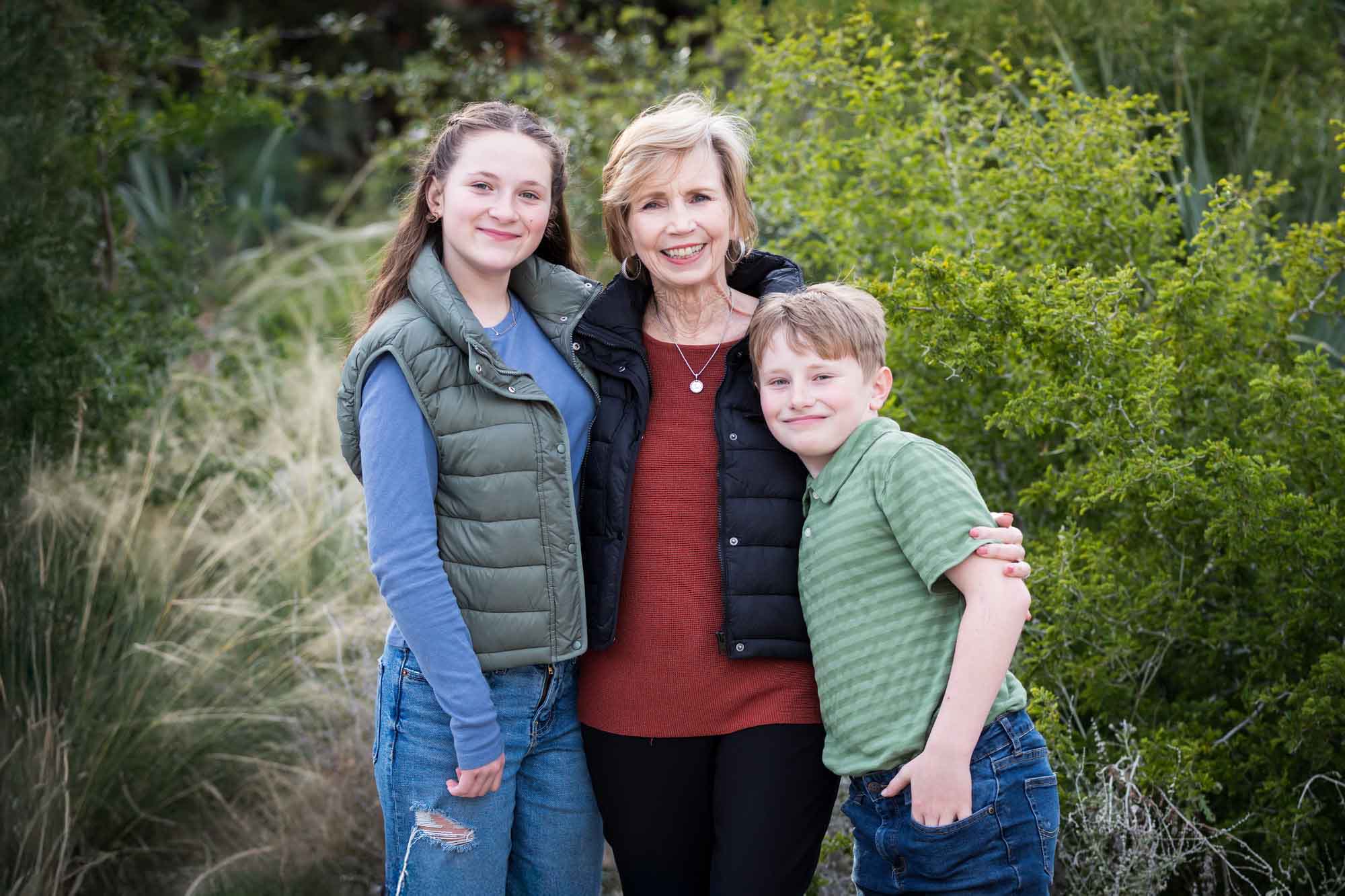 Grandmother hugging granddaughter and grandson in front of green bushes during a Hemisfair Park family portrait session
