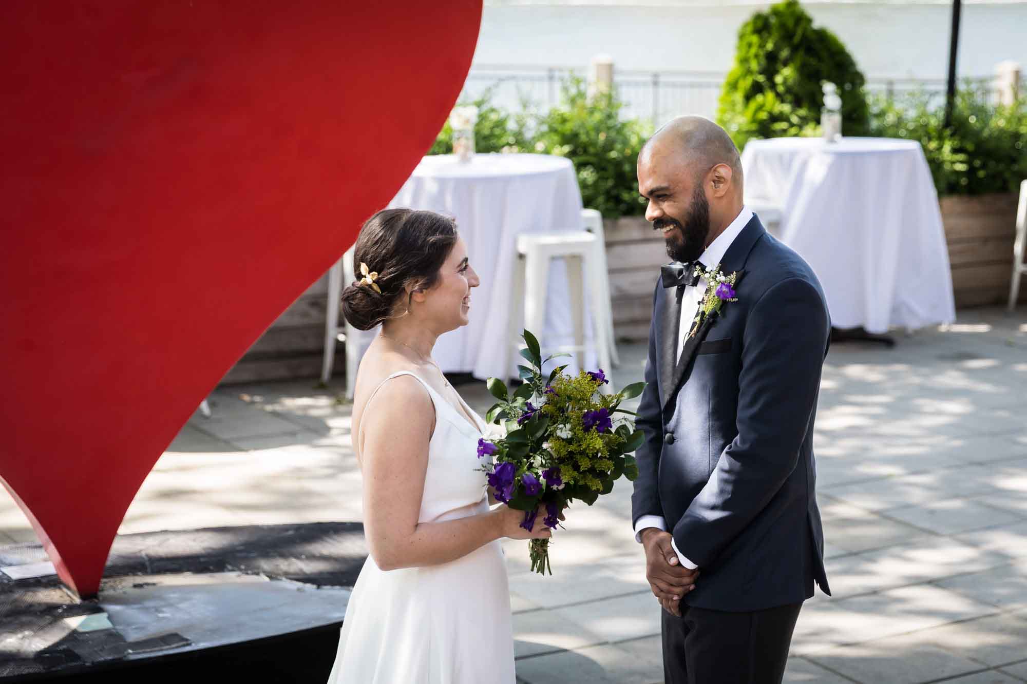 Bride and groom standing on patio in front of large red heart during first look for article on pros and cons of first look