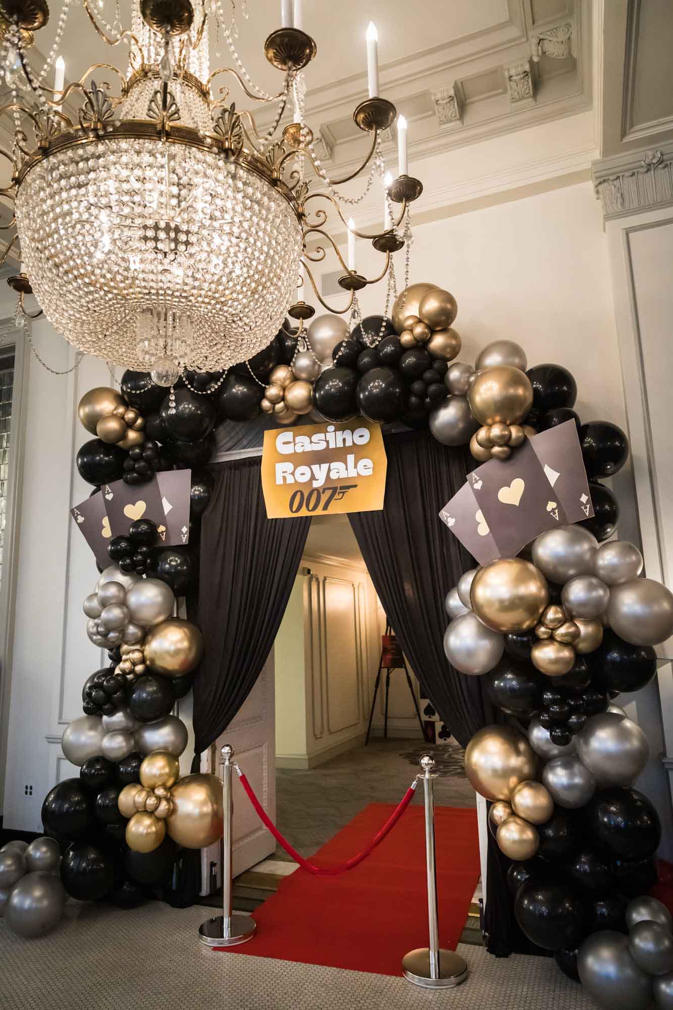 Balloon archway decorated in black, gold, and silver Casino Royale theme underneath large chandelier and red carpet going through sculpture