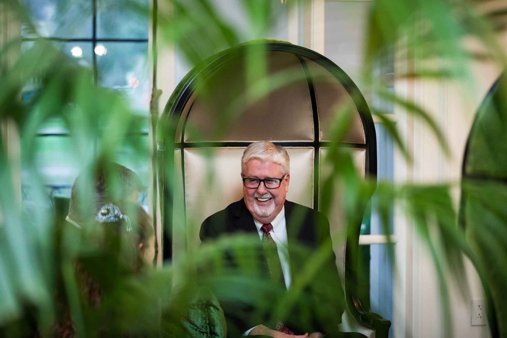 Older man wearing glasses and laughing while wearing black jacket and tie as viewed through a green plant for an article on a St. Anthony Hotel corporate event