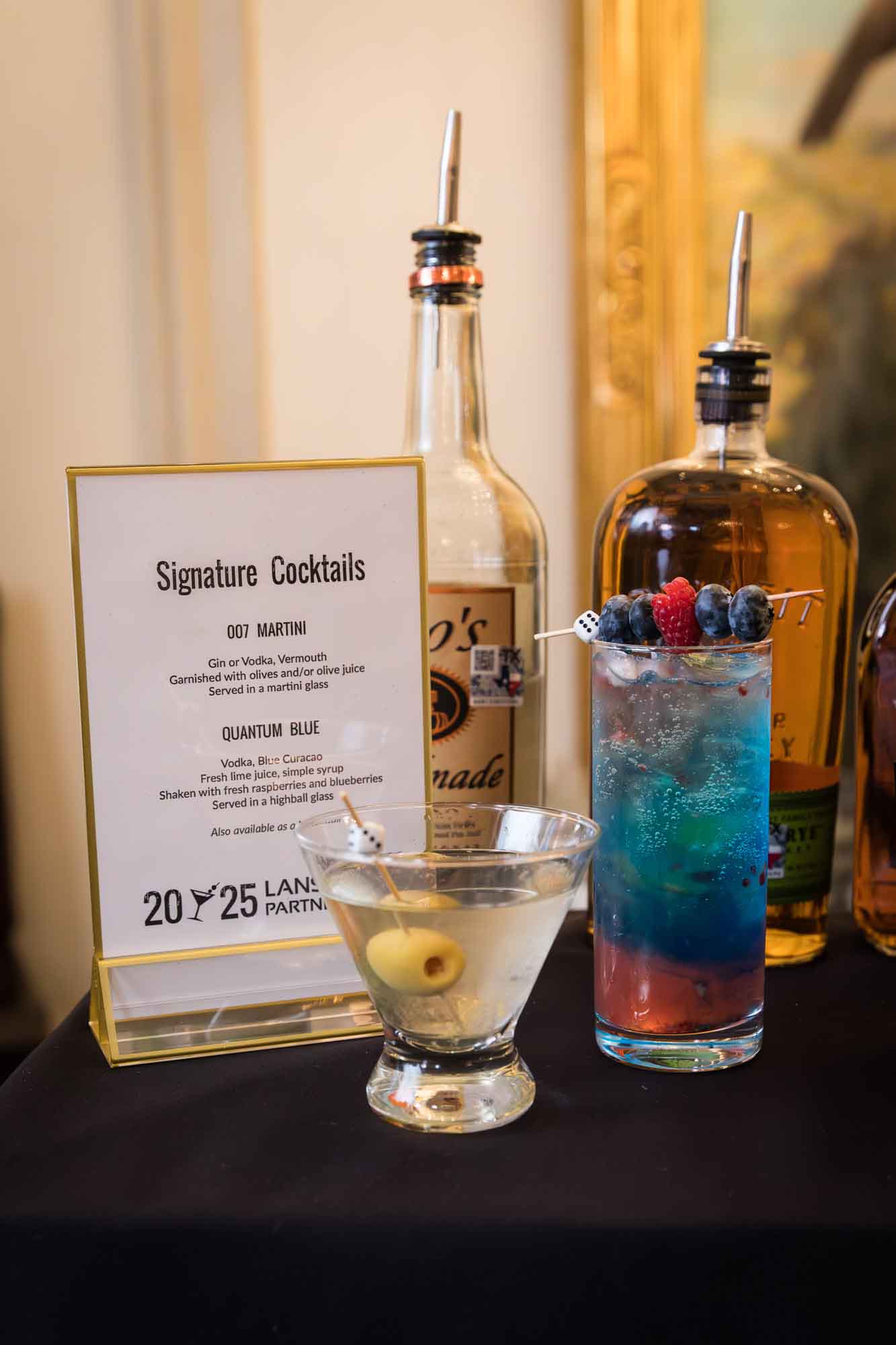 A martini glass with an olive and a tall blue cocktail with berries on top in front of two bottles of liquor and a sign for an article on a St. Anthony Hotel corporate event
