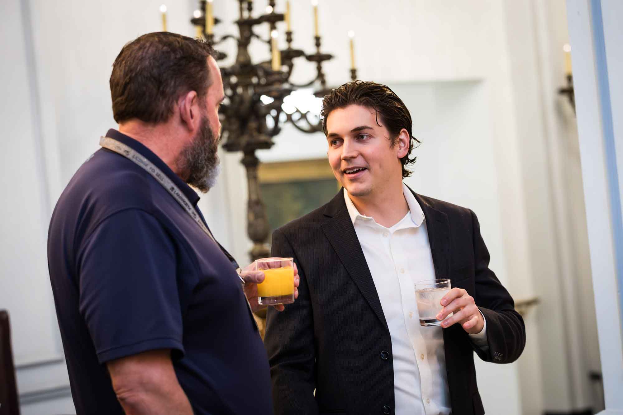 Two men holding drinks and speaking in front of chandelier for an article on a St. Anthony Hotel corporate event