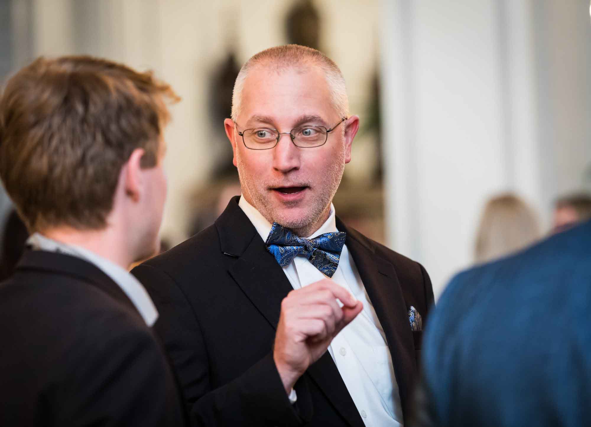 Man wearing glasses and a blue paisley bow tie speaking with hand raised for an article on a St. Anthony Hotel corporate event