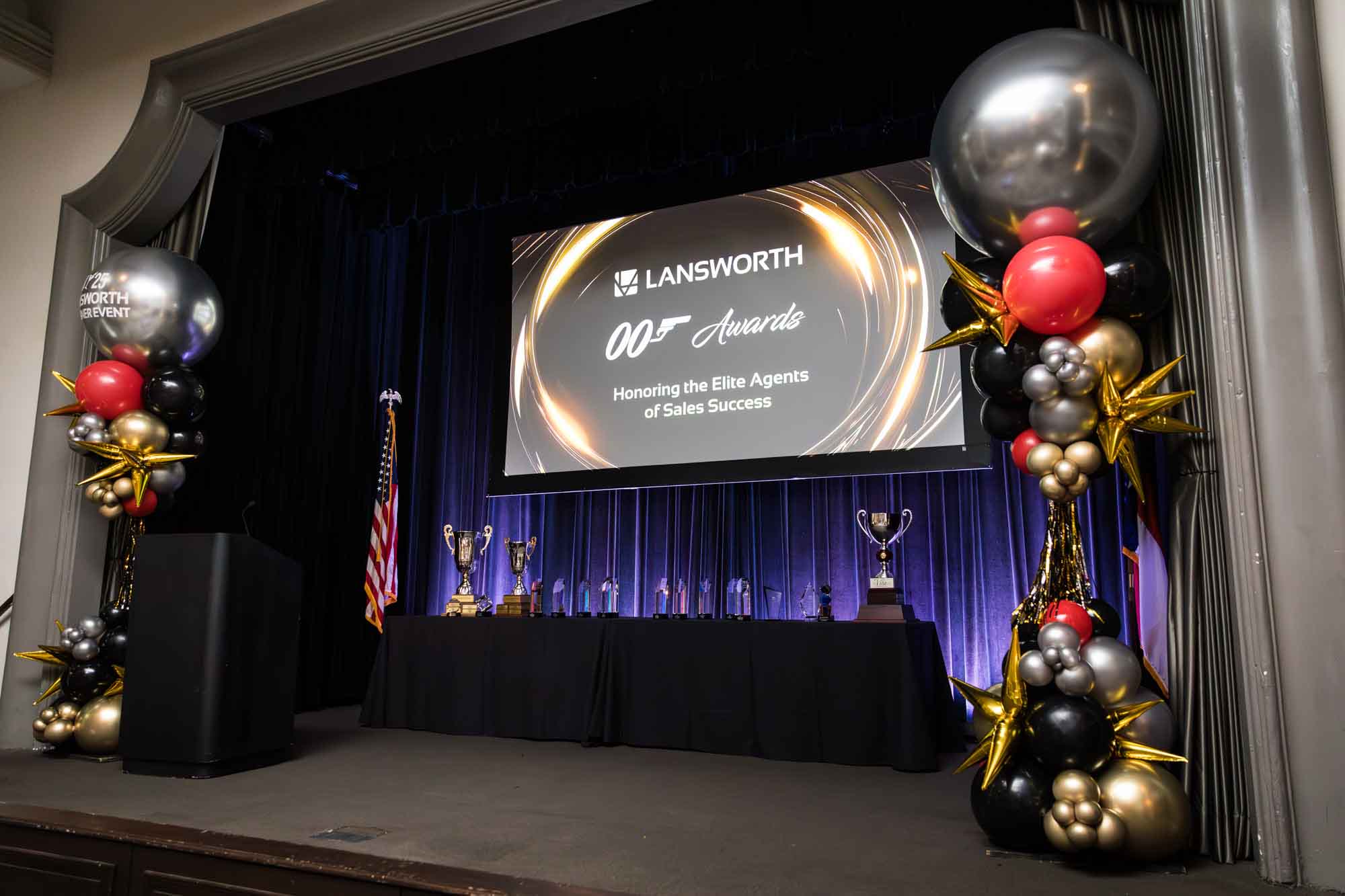 Stage for awards ceremony with balloon sculptures and table with awards, screen, and American flag