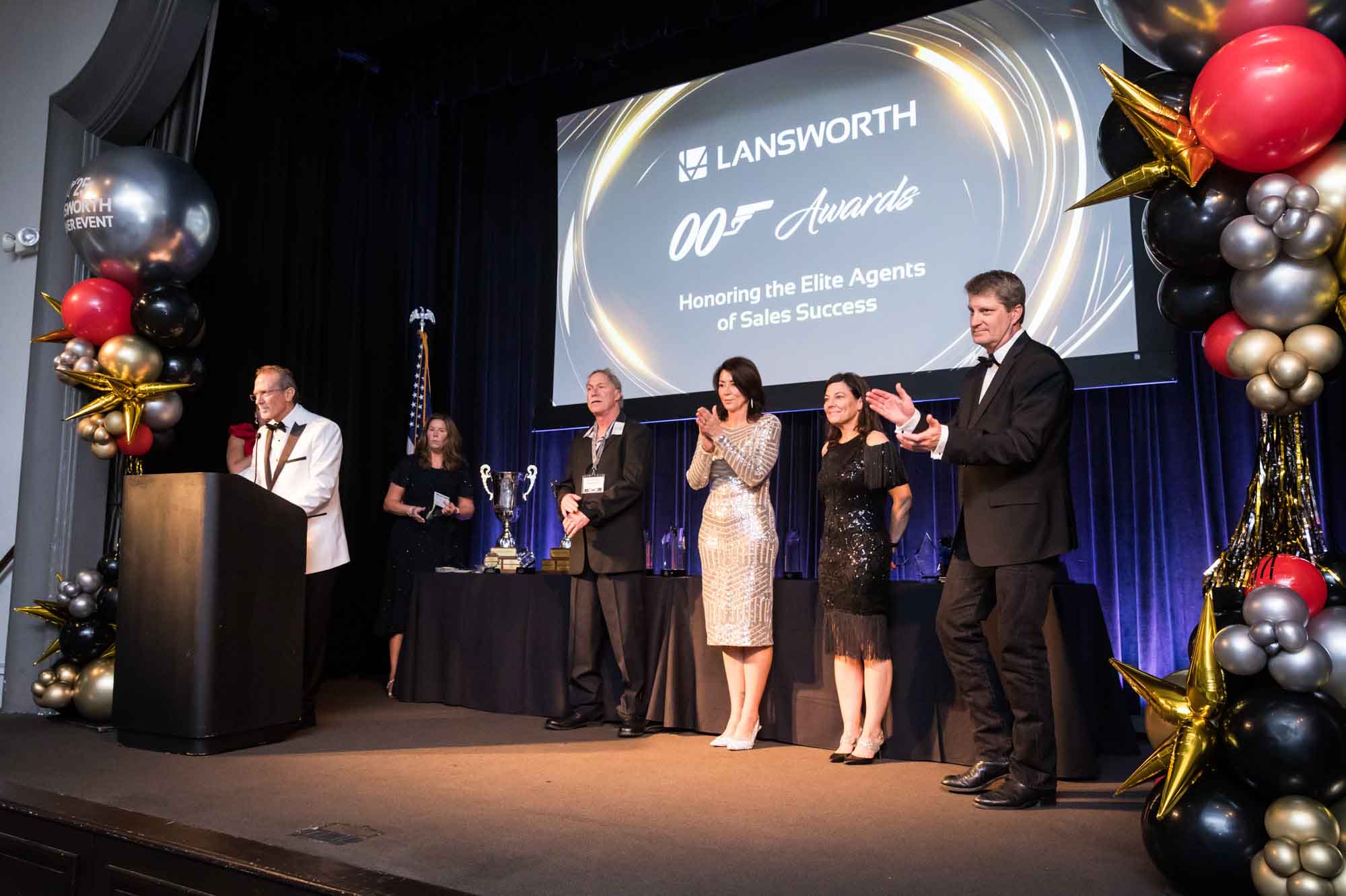 Man in white tuxedo jacket speaking at podium speaking in front of line of people with balloon sculptures and screen in background for an article on a St. Anthony Hotel corporate event