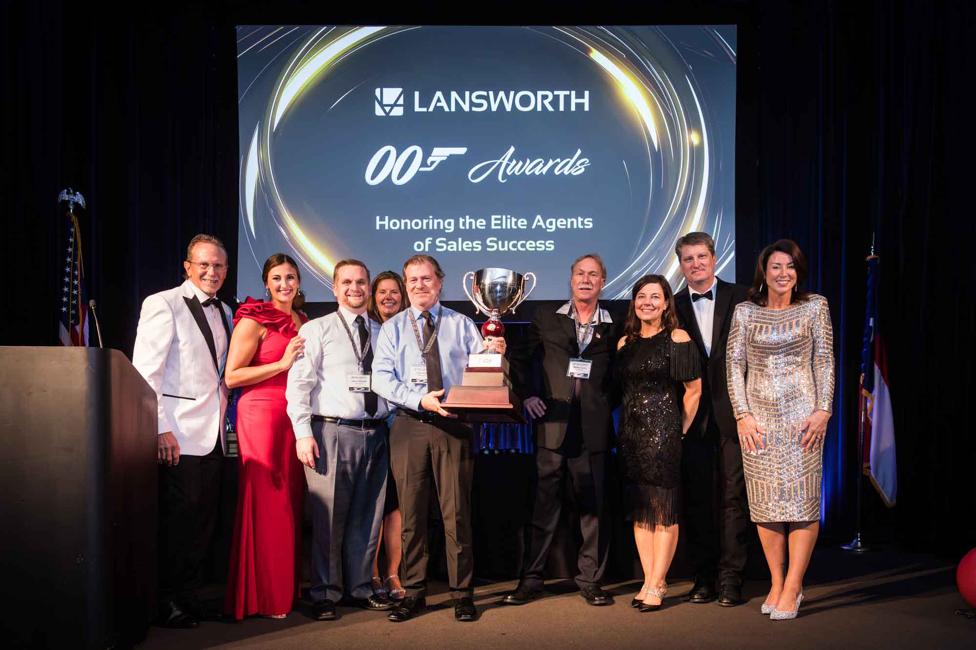 Line of award winners holding up large silver award cup in front of screen for an article on a St. Anthony Hotel corporate event