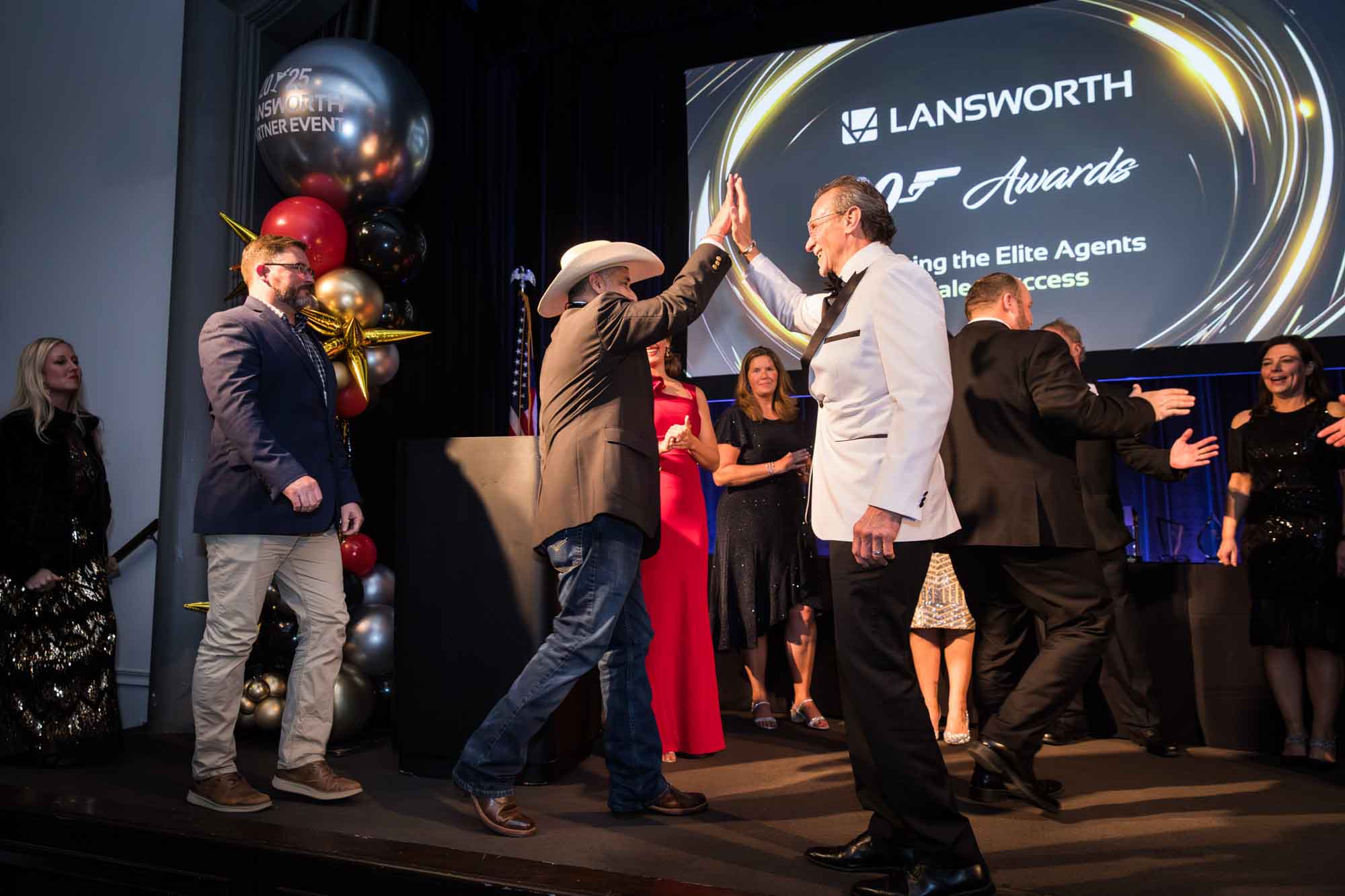 Man in white tuxedo jacket high-fiving man wearing cowboy hat as he walks across the stage for an article on a St. Anthony Hotel corporate event