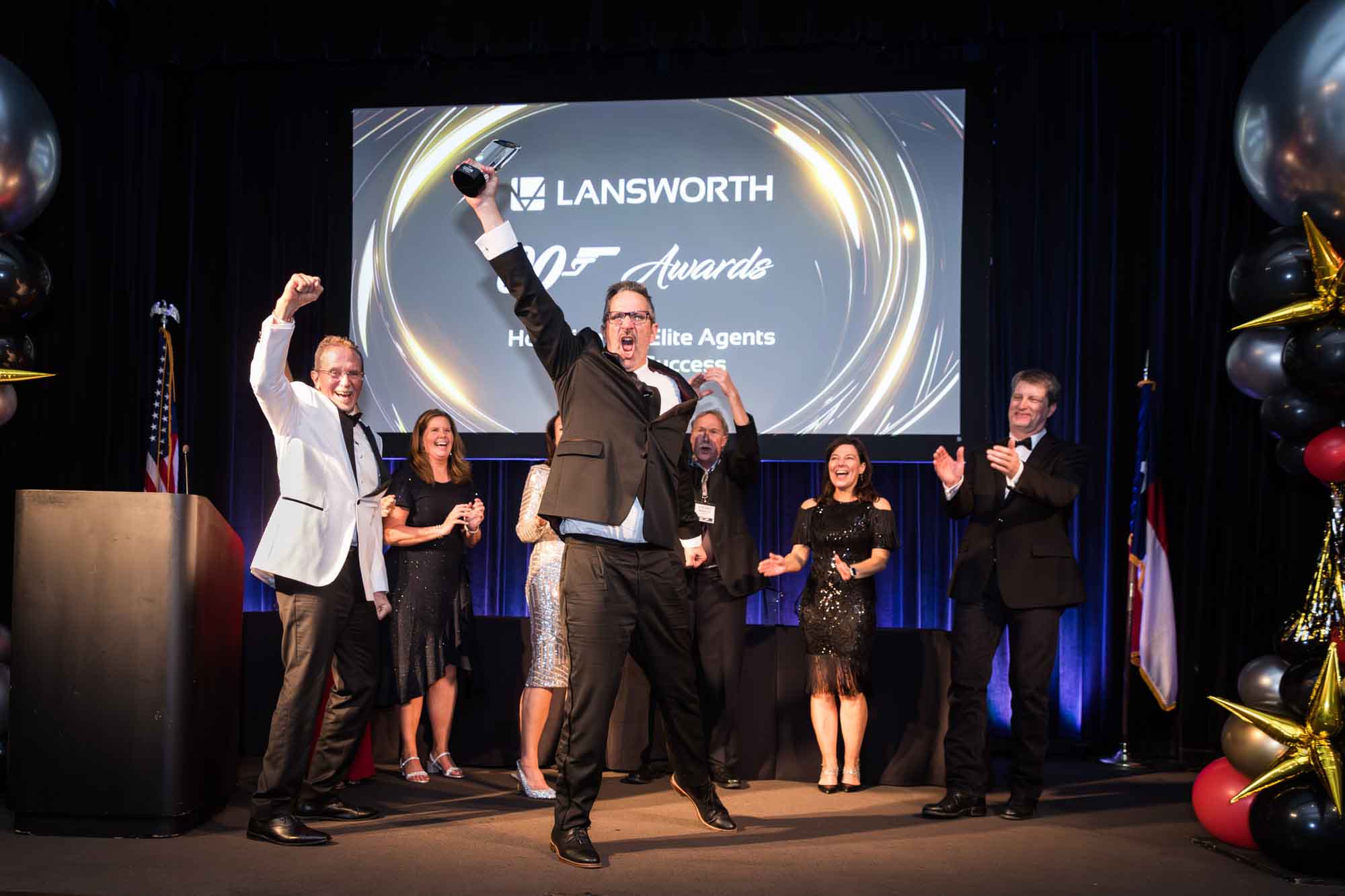 Man wearing black suit excitedly holding up award in the air in front of line of clapping people and screen in background for an article on a St. Anthony Hotel corporate event