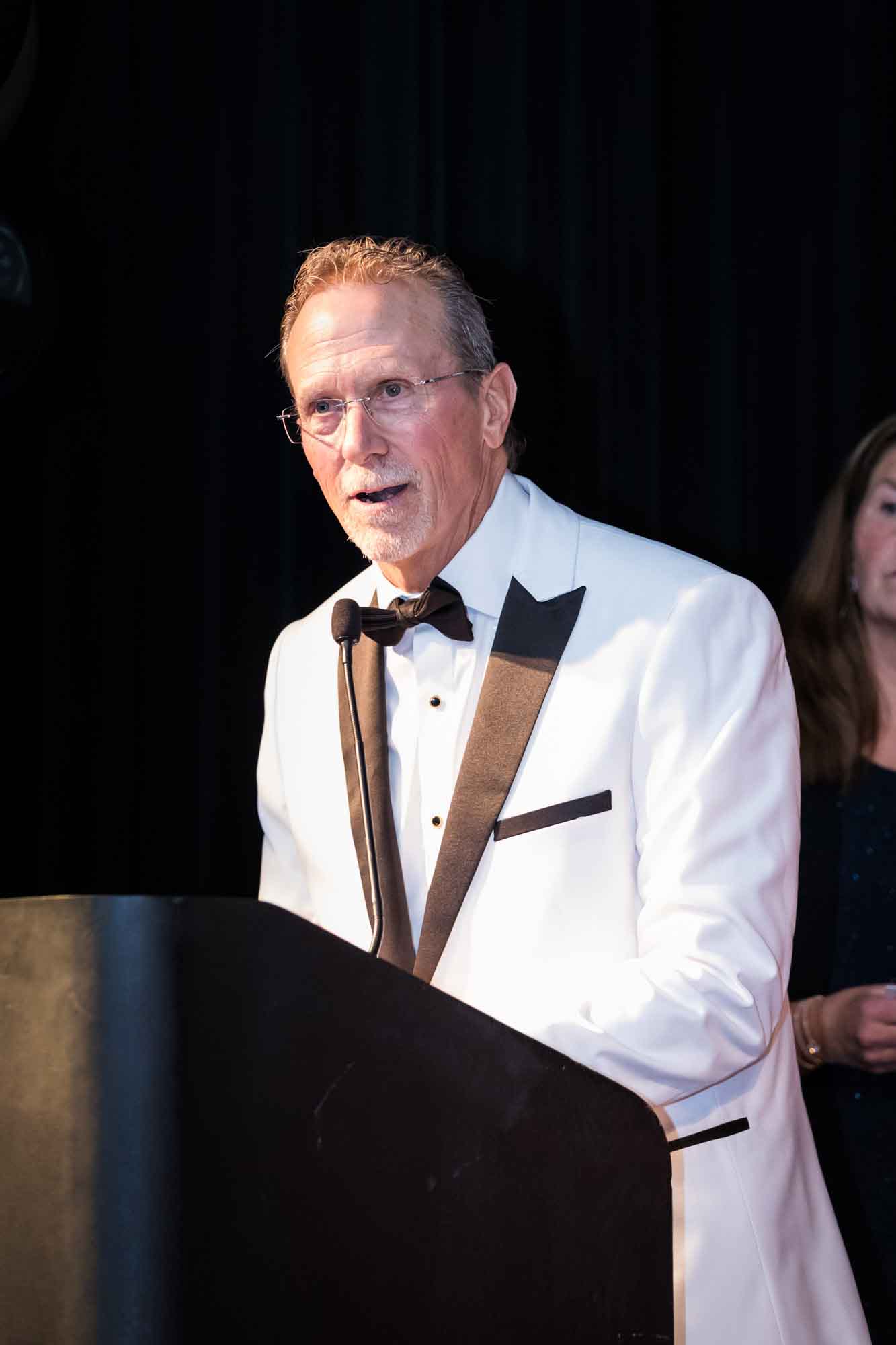 Man wearing white tuxedo jacket and glasses speaking at podium for an article on a St. Anthony Hotel corporate event
