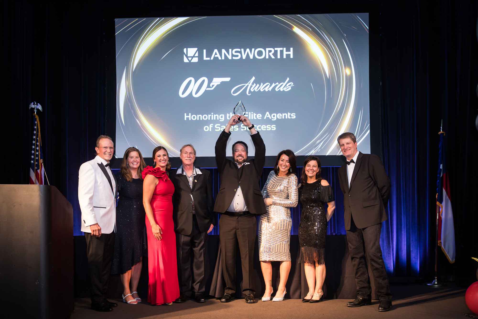 Line of award winners with man holding up glass award in front of screen for an article on a St. Anthony Hotel corporate event