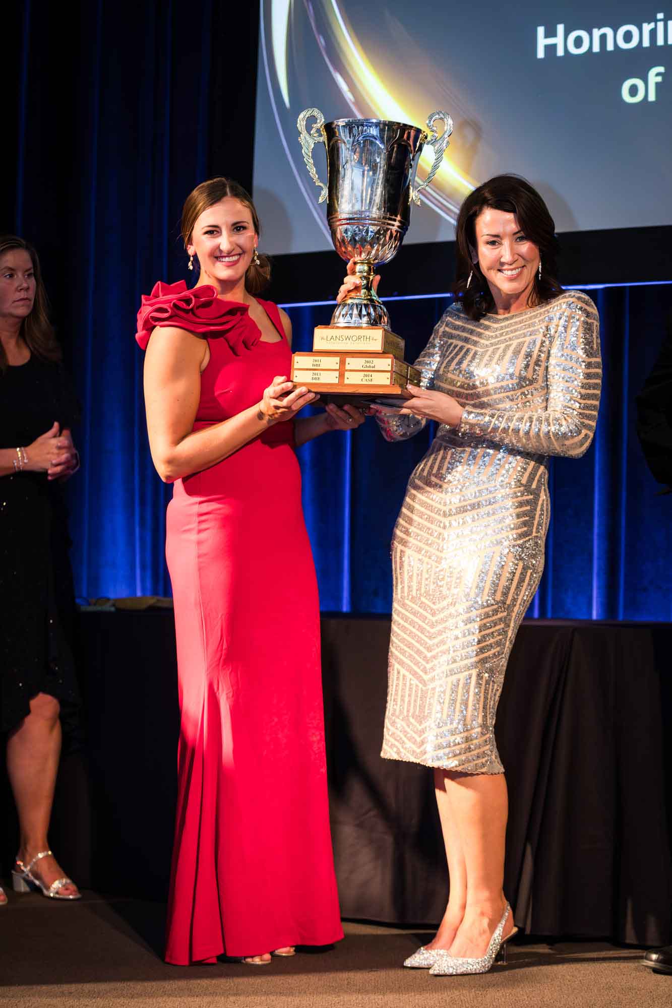 Woman wearing long red dress and woman wearing silver sequined dress holding large silver award cup for an article on a St. Anthony Hotel corporate event