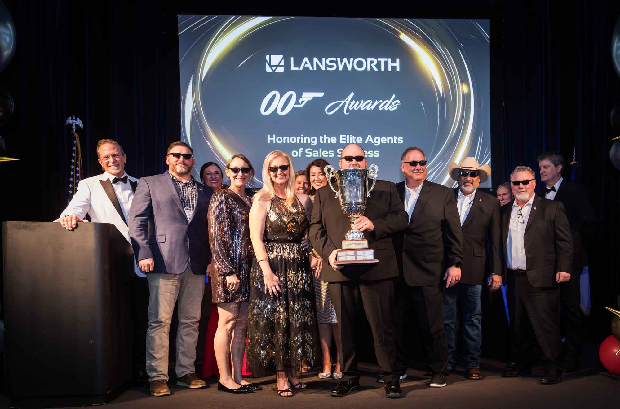 Row of award winners wearing black sunglasses holding large silver award in front of screen for an article on a St. Anthony Hotel corporate event