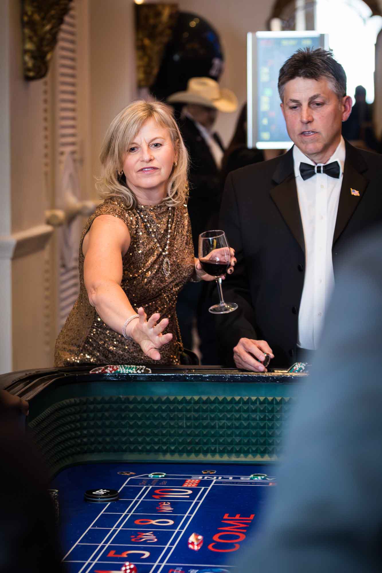 Woman wearing gold sequin dress holding wine glass throwing dice at a craps table throwing dice in front of man wearing bow tie for an article on a St. Anthony Hotel corporate event