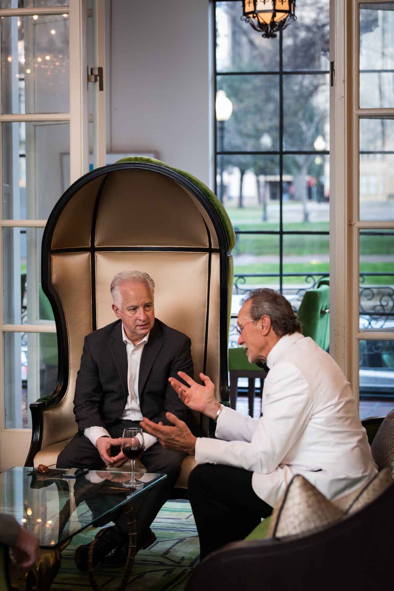 Two older men seated while chatting in front of a window for an article on a St. Anthony Hotel corporate event