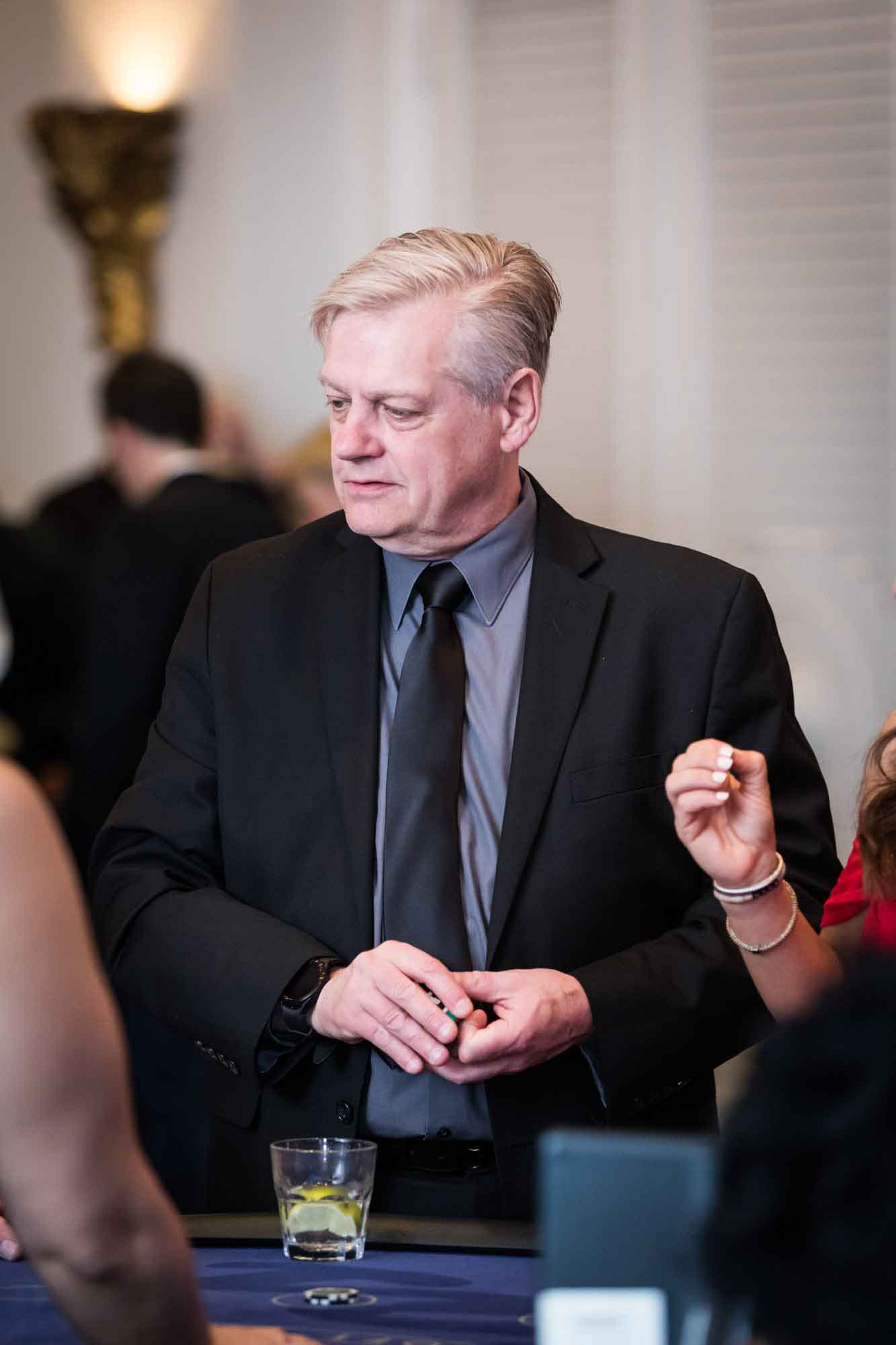 Man wearing a black jacket and tie standing in front of a card table with a glass in front of him for an article on a St. Anthony Hotel corporate event