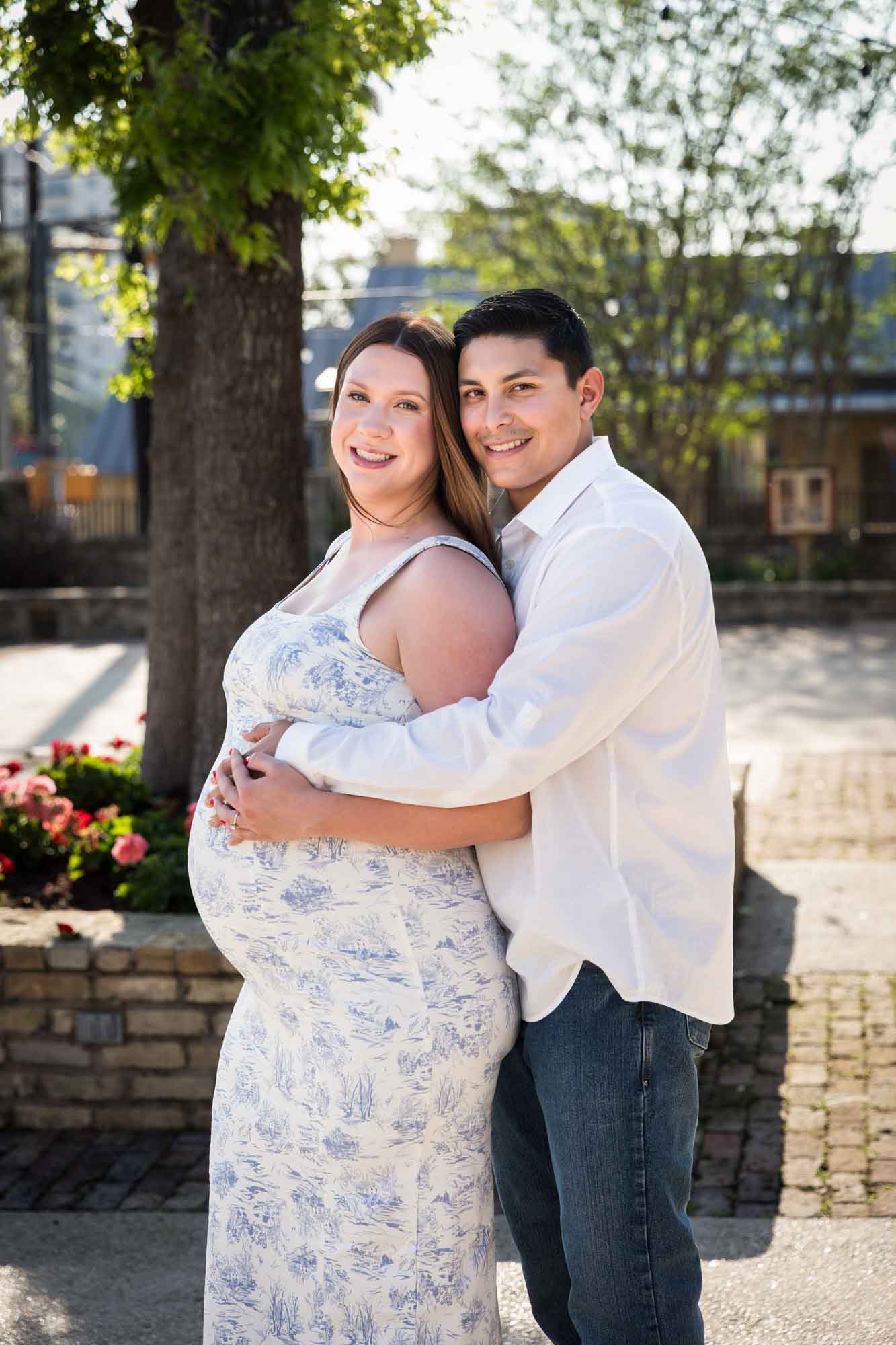 Pregnant woman and man hug in a patio in front of a tree during a La Villita engagement portrait session