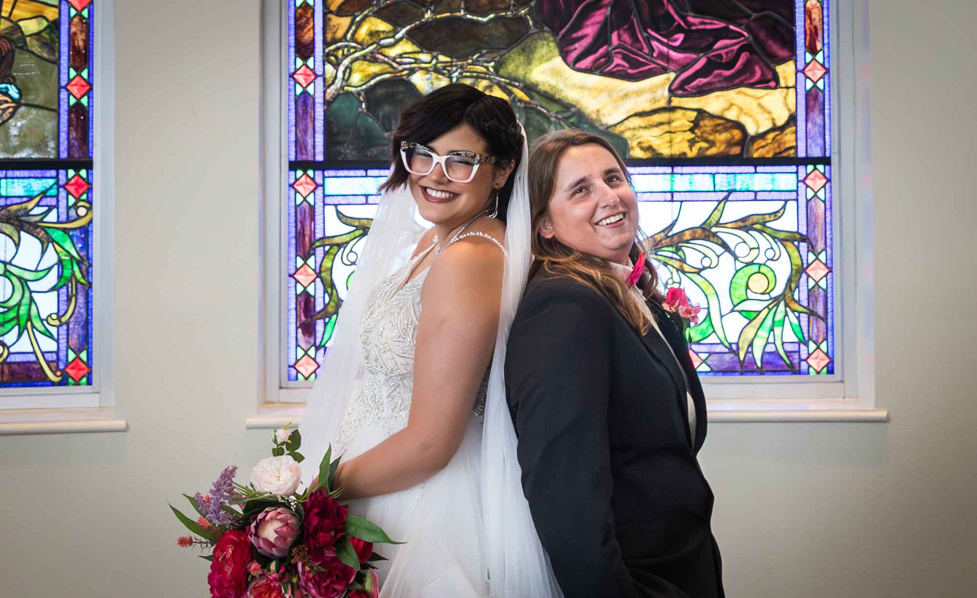 Bride and groom standing back to back with stained glass windows in background in Vada Hagee Prayer Chapel