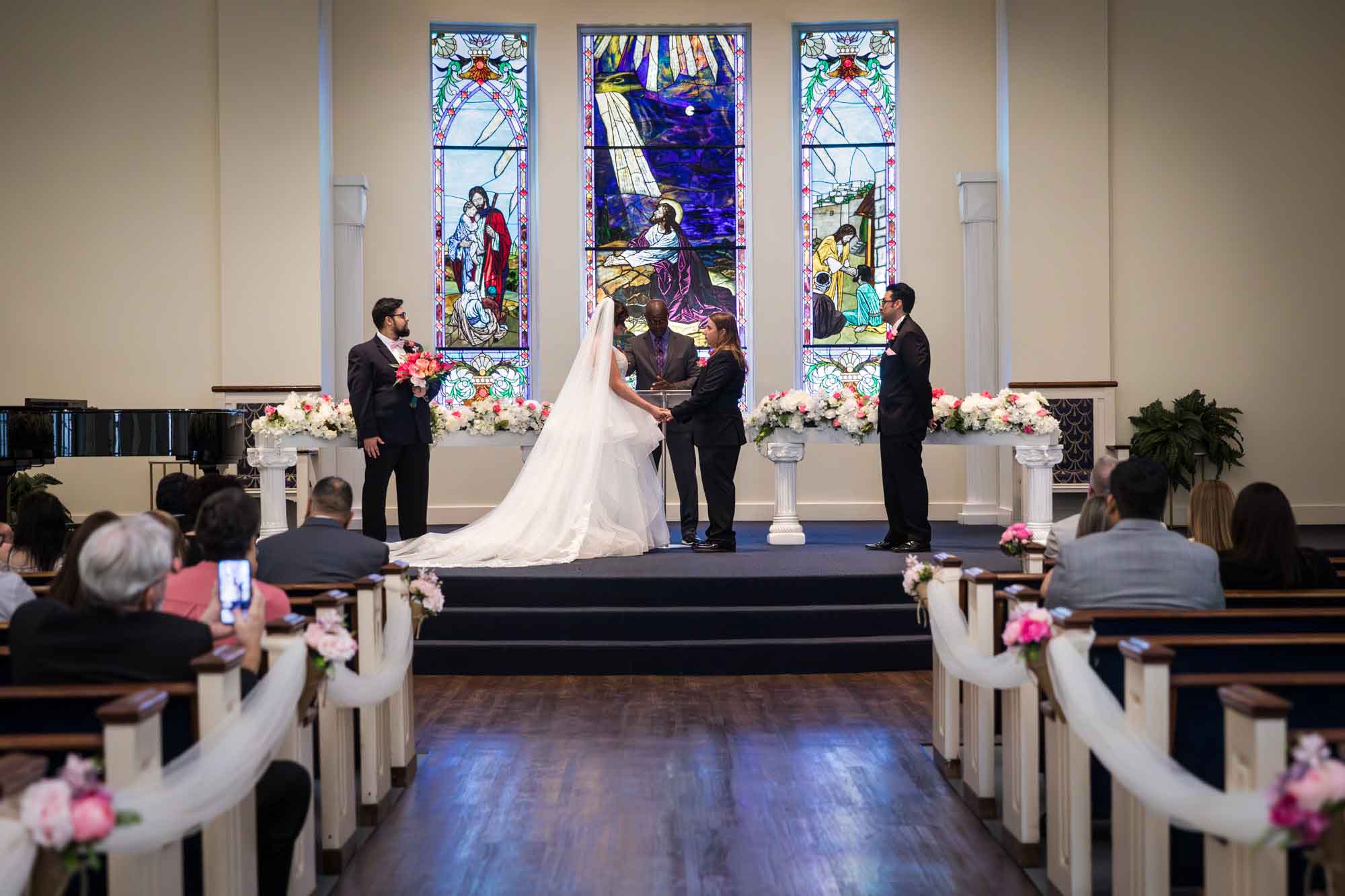 Cornerstone Church wedding photos of bride and groom holding hands with two groomsmen on either side and stained glass windows in background and guests seated in pews