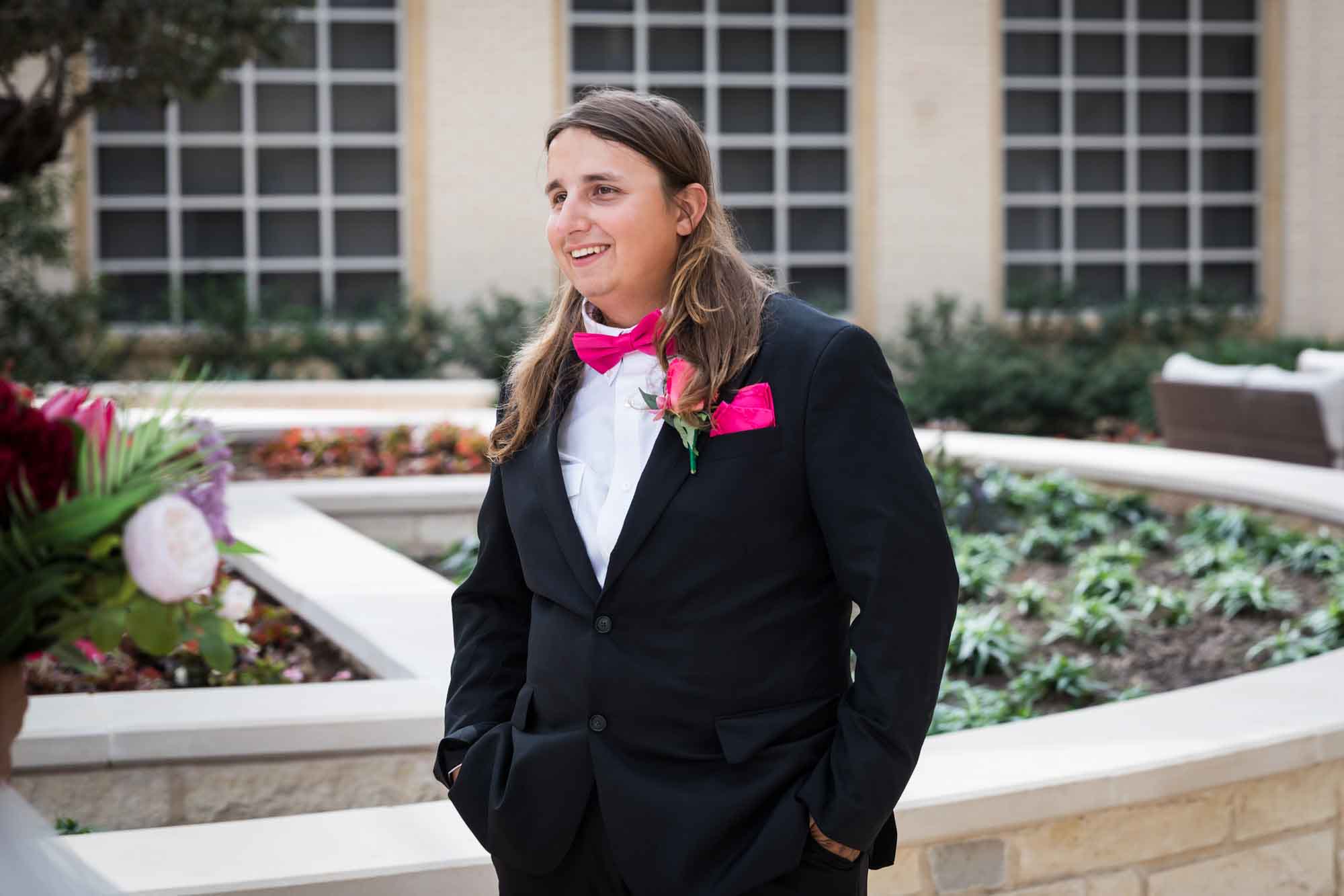 Cornerstone Church wedding photos of groom wearing black suit and pink bow tie standing in front of large planter in courtyard