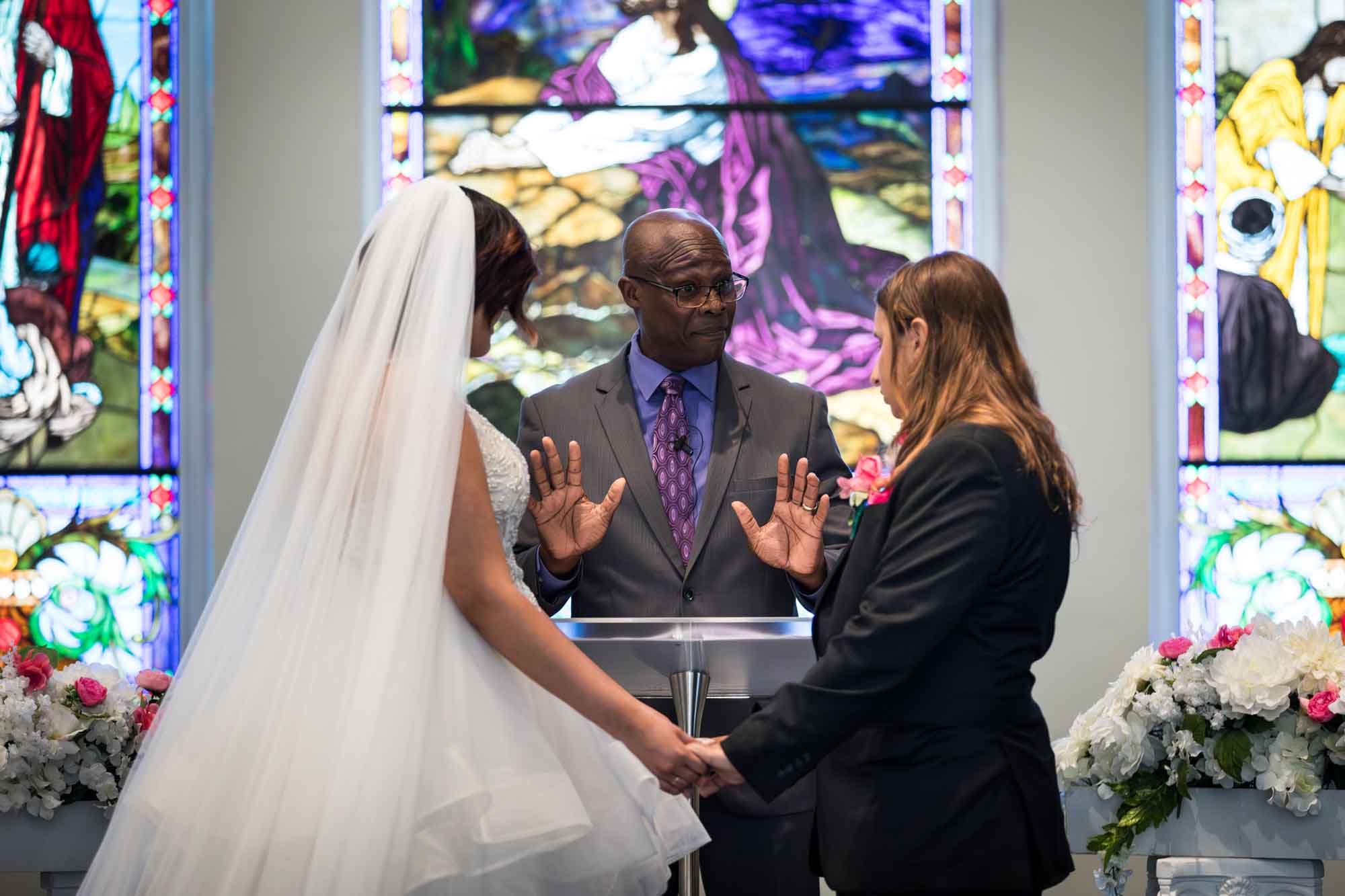 Cornerstone Church wedding photos of bride and groom holding hands in front of African American officiant with stained glass windows in background