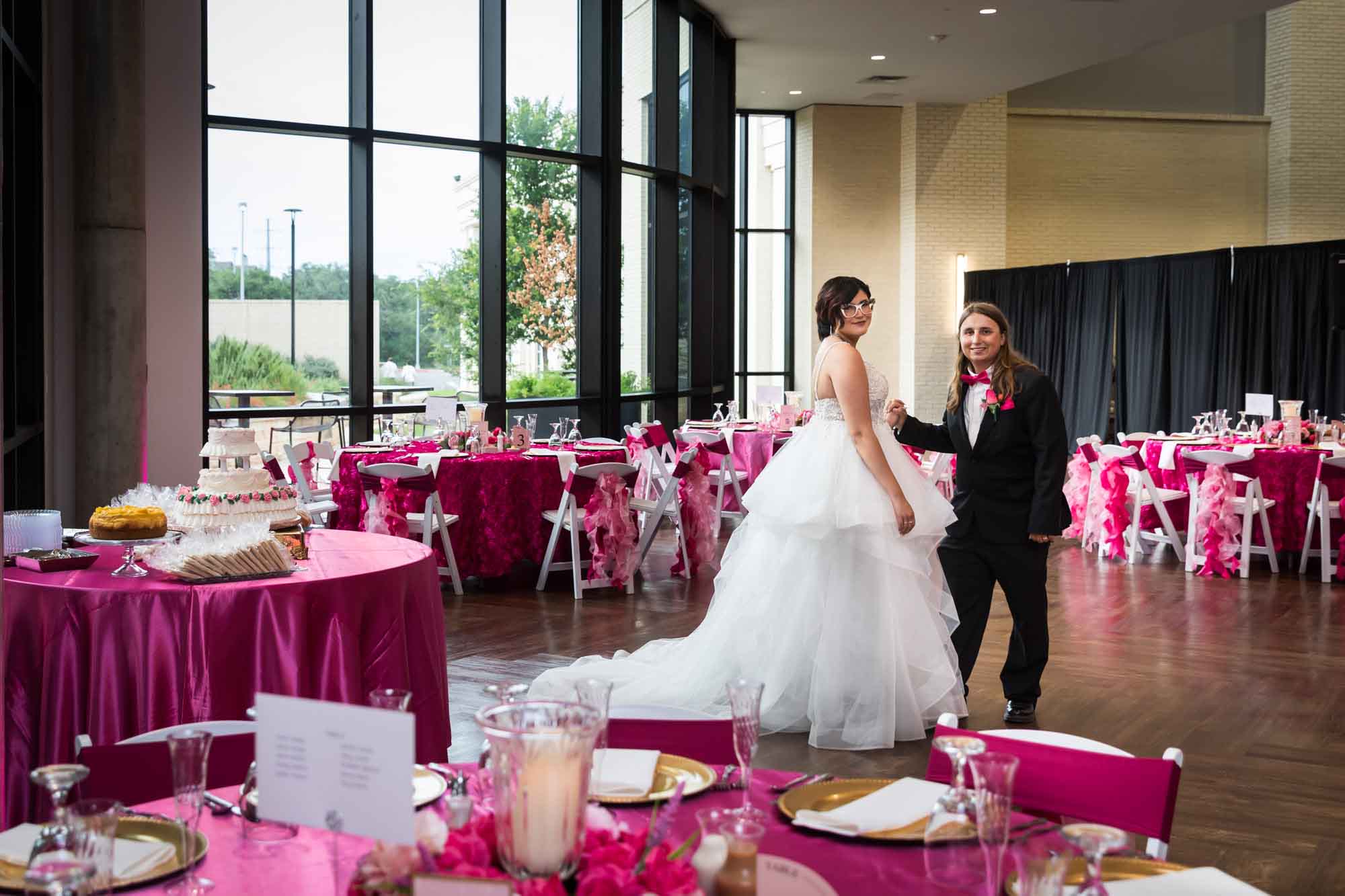 Bride and groom standing in the middle of a wedding reception room filled with tables decorated with pink tablecloths at Cornerstone Church